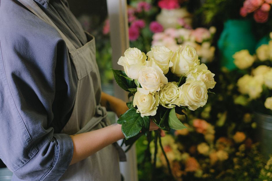 A person in a gray apron holds a bouquet of fresh, cream-colored roses with lush green leaves, arranging the floral gift inside a flower shop. The roses are in full bloom, with delicate, velvety petal