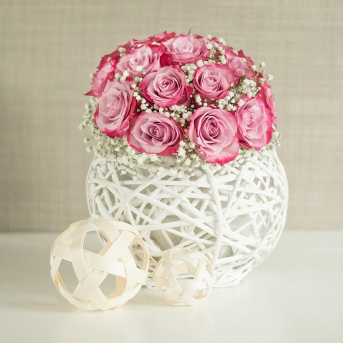 A serene interior scene features a rounded bouquet of pink roses and white baby's breath arranged in a distinctive spherical white vase, flanked by two woven decorative spheres, creating a refined, contemporary look that feels typical of stylish homes around Brent Cross and nearby Golders Green. The bouquet is dense and carefully balanced, with each rose placed so that only the open blooms are visible, forming a gentle dome shape. The roses themselves show a beautiful two-toned effect: at their centers, the petals are a soft, almost pastel blush, while toward the edges the pink deepens into a more vivid, raspberry or fuchsia shade, giving the arrangement both softness and vibrancy. Scattered between the roses, clusters of gypsophila add a lace-like lightness, their tiny white blossoms and fine stems breaking up the solid masses of color and enhancing the romantic mood. The flowers rest in a round white container designed from overlapping strips that look like painted rattan or wicker, the open, airy pattern creating a sense of handcrafted artistry. Light filters through the gaps in the weave, casting subtle patterns of light and shadow on the smooth white tabletop below. To the left of the vase, two sculptural spheres made of thin, pale wooden or natural-fiber strips sit close together, echoing the vase's texture and adding a touch of minimalist, organic decor. The backdrop is a softly textured wall in warm beige or cream, unobtrusive yet elegant, which allows the pink and white tones to stand out clearly. The lighting is bright but evenly diffused, reminiscent of daylight on a clear morning near Brent Cross Shopping Centre, lending the arrangement a fresh, welcoming feel. Overall, the composition suggests a thoughtfully styled floral centerpiece, ideal for an intimate celebration, a welcoming hallway display, or inspiration for a locally crafted arrangement.