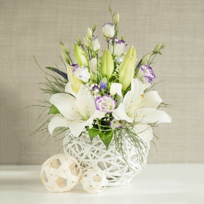 A refined centerpiece designed for a wedding reception or special event is arranged in a rounded white vase with a striking open-weave design, reminiscent of delicate wickerwork. The vase rests on a clean white tabletop, set against a soft, neutral beige wall with a faint horizontal linen effect that adds gentle visual interest without stealing attention from the flowers. The arrangement is dominated by large, white lilies, some fully open with broad, slightly recurved petals and visible stamens tinged with pollen, others presented as elongated green buds that rise above the main body of the bouquet. Interlaced with the lilies are generous clusters of white lisianthus, whose layered petals create a rose-like fullness, many of them edged or gently brushed with pale lavender-purple tones. Small, deeper purple filler blooms and clouds of tiny white florets nestle between the larger flowers, creating depth and a soft, romantic haze. The greenery is abundant yet carefully balanced, including feathery, almost fern-like sprays and smooth, broader leaves that spill lightly over the rim, giving the design a natural, garden-inspired flow. Beside the vase, two decorative spheres of different sizes, crafted from a light, woven material, rest on the tabletop and echo the vase's lattice motif, bringing a cohesive, thoughtfully styled look. The bright, even light highlights each petal, leaf, and woven strand, accentuating the contrast between crisp white, tender green, and gentle purples. This arrangement conveys an atmosphere of purity, serenity, and quiet joy, ideal for adorning wedding tables in and around Brent Cross, from elegant venues near the shopping centre to intimate gatherings in homes along Hendon Lane or the leafy streets towards Finchley.