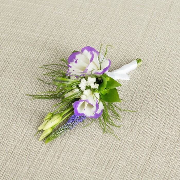 This image shows a meticulously crafted floral boutonniere resting on a softly textured, light beige fabric, the sort of neutral backdrop that resembles natural linen and lets the colours of the flowers quietly glow. At the centre, two fully opened lisianthus flowers draw the eye with their smooth white petals and distinctly ruffled edges, each rim carefully brushed in a rich, vibrant purple that creates a flowing, bi-colour halo effect. Tucked around these focal blooms are tiny clusters of white filler flowers, similar to baby's breath, that add airy volume and soften the composition like a delicate veil. A slender spike of grape-hyacinth-style muscari rises alongside them, its tightly packed, bell-shaped blossoms a deep, inky purple, introducing a vertical element and a slightly more intense hue. Pale green buds, not yet unfurled, sit among the flowers, suggesting a living moment paused just before full bloom, as though the design has come directly from a fresh delivery to a Brent Cross home or wedding venue. Around the flowers, a mix of greenery provides contrast: broader, deeper green leaves give structure at the base, while feathery, fern-like foliage reaches lightly outward, creating a sense of gentle movement. The stems are carefully aligned and wrapped in a crisp band of white satin ribbon, smooth and slightly luminous, tied off neatly to mark this as a finished piece ready for a lapel. The lighting is soft and even, emphasising the fresh textures and classic palette, making this boutonniere ideal for a groom, groomsman, or formal guest attending an elegant event anywhere around Brent Cross and nearby North London suburbs.