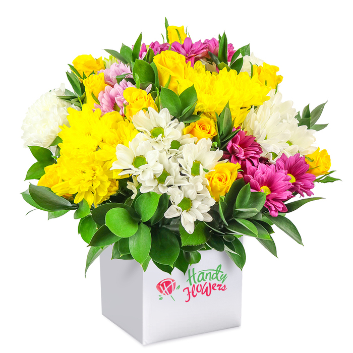 Set against a clean white backdrop, this densely packed, dome-shaped bouquet in a white square box captures the eye with its harmony of yellow, white, and vivid magenta tones. At first glance, the sunny yellow chrysanthemums dominate, their many soft petals forming large, fluffy blooms that give the arrangement a warm, inviting heart. Around them, clusters of yellow spray roses at different stages of opening-from tight buds to partially unfurled blossoms-add detail and a sense of gentle movement. Woven into this golden base are clusters of white chrysanthemums and daisy-like flowers with white petals and fresh green centres, creating crisp highlights and visual breathing space, reminiscent of bright daylight over nearby Brent Cross streets and gardens. The boldest accents come from the fuchsia and deep magenta chrysanthemums scattered throughout, their layered petals sometimes flushed with lighter pink, adding depth and a slightly romantic feeling to the otherwise cheery palette. Deep green, glossy leaves surround the lower edge and peek between the flowers, providing a lush, structural frame that reinforces the dome shape and keeps each colour distinct. The container is a minimalist white cube, its straight lines contrasting with the bouquet's rounded form and giving the design a contemporary, gift-ready character, perfect for placing on a dining table in a Brent Cross home or on a desk in an office near the North Circular. Together, the colours, textures, and careful composition suggest a fresh, affordable arrangement suited to birthdays, thank-you gestures, or brightening any everyday moment with local delivery convenience.