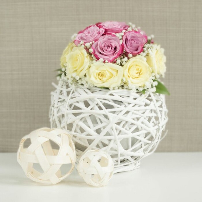 This image presents a carefully styled rose centerpiece, ideal for wedding tables or intimate celebrations across Brent Cross and nearby Hampstead Garden Suburb. A spherical bouquet of roses crowns a white wicker globe vase, the blooms arranged in a close and harmonious dome. The colour palette is soft and balanced: half the roses are a gentle, pastel pink with touches of lavender and deeper fuchsia in the inner petals, while the other half glow with creamy yellow and ivory tones. The petals show a range of openness-from tight, promising buds to fully unfurled faces, revealing the curled spiral at the heart of each flower. Between the roses, fine sprigs of white baby's breath appear in small clouds, adding both volume and a delicate texture that suggests bridal lace. Supporting this bouquet is a distinctive vase shaped like a sphere, woven from slender white branches or wicker that criss-cross into an open, lattice-like structure. Through the spaces in the weave, slight shadows and the darker stems of the roses can be seen, creating depth and contrast against the bright white exterior. The vase sits on a smooth, white tabletop that feels calm and uncluttered. To the left, two decorative spheres crafted from light natural wood or rattan rest on the same surface, their open-weave design echoing the globe vase but in a warmer, more rustic tone. The background is a soft, neutral beige or light taupe wall, subtly textured and gently lit, allowing the blush and cream hues of the flowers to stand out. The whole effect is one of understated sophistication and warmth, reminiscent of a beautifully set table in a Brent Cross apartment awaiting guests for a special celebration.