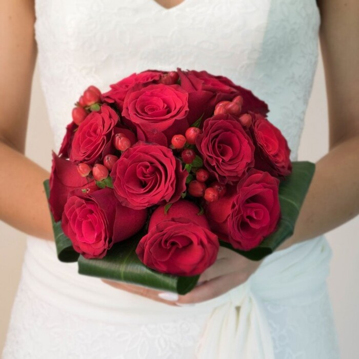 A masterfully composed close-up reveals a Brent Cross bride in a pristine white dress, her hands tenderly embracing a magnificent bridal bouquet. The luxurious arrangement is a heartfelt celebration of love, its compact, domed structure showcasing countless velvety, deep red roses-each bloom nestled closely to its neighbour in a display of unity and elegance. Sprinkled amid the roses, gleaming red hypericum berries add a dynamic touch, their festive energy reminiscent of vibrant gatherings beside the River Brent. Encasing the bouquet, broad, dark green leaves have been carefully crafted into a structured collar, offering striking visual contrast and a touch of contemporary flair that echoes local Brent Cross green spaces. The bride's neatly manicured hands suggest quiet excitement, while her gown's intricate lace details and gently draped fabric provide a luminous, romantic setting. Bathed in natural, flattering light, every element-from the rose petals' silky texture to the cooling green of the foliage-blends seamlessly, testifying to the artistry of Brent Cross Flowers. An ideal bouquet for wedding ceremonies, reception décor, or local bridal features, this composition fuses classic beauty with a distinctly Brent Cross sense of place.