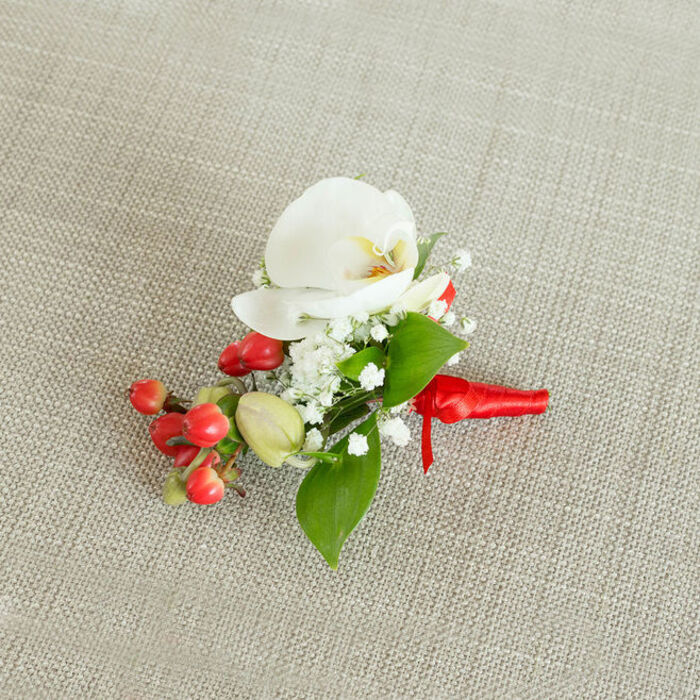 This detailed image shows a refined floral boutonniere or small corsage laid carefully on a light beige, textured textile that resembles natural linen, its woven threads visible and softly catching the light like a table runner at a Brent Cross wedding breakfast. At the centre of the arrangement is a single white Phalaenopsis orchid, its generous, rounded petals elegantly fanned out in a calm, symmetrical shape. The orchid's throat glows with subtle tones of creamy yellow and muted orange, adding an inner warmth that contrasts gently with the cool whiteness of the outer petals. Around this focal bloom, clouds of tiny white baby's breath blossoms expand the composition, each miniature flower adding airiness and a sense of delicate movement. To one side of the orchid, a tight cluster of glossy, round red berries offers a striking burst of colour, their rich, festive hue reminiscent of holly or winter hypericum often used for seasonal ceremonies near Hendon Way and the wider Brent Cross community. Among these berries, a few smaller, paler green buds can be seen, hinting at new growth and adding subtle variation in size and shade. A selection of smooth green leaves, some elongated, some more oval, thread in and out of the piece, giving it structure and a fresh, garden-grown feel. The stems are bound neatly with a vivid red satin ribbon, wrapped with precision and tied in a compact knot, its short tails resting gracefully along the neutral fabric. The soft, natural light reveals every texture-from the orchid's satiny surface to the berries' glossy sheen-creating a sense of quiet celebration. This boutonniere feels ideal for a groom, best man, or prom guest attending a special event around Brent Cross, combining classic elegance with a touch of seasonal charm.