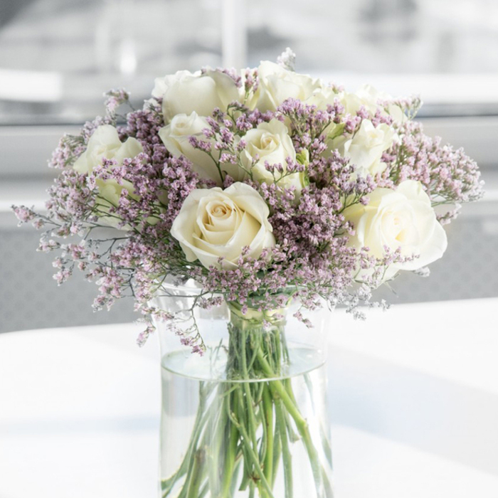In this image, a refined bouquet of creamy white roses and lavender filler flowers stands poised in a tall clear glass vase, exuding a sense of serene dignity well suited to sympathy occasions in Brent Cross. The roses are generously proportioned, each bloom fully open and revealing a complex swirl of soft petals that spiral inward. Their colour is a gentle progression from white along the outer petals to a mellow ivory at the centre, evoking purity paired with quiet warmth. Threaded among these larger blooms are abundant sprigs of small, cloud-like blossoms in soft lavender, lilac, and pale pink, likely statice or limonium, adding an airy, almost ethereal quality. These tiny flowers form a delicate halo around the roses, their fine texture contrasting with the roses' velvety surfaces and giving the arrangement a feeling of lightness despite its fullness. The cylindrical glass vase allows a clear view of the long, green stems, some slightly angling, others almost straight, all immersed in fresh, clear water that catches the ambient light. This transparent base grounds the bouquet with simple elegance, much like the understated yet contemporary feel of homes around Brent Cross and nearby Golders Green. The vase is placed on a pristine white surface, subtly reflecting the lower portion of the glass and brightening the composition from below. Behind the bouquet, the background is softly blurred into pale greys and whites, suggesting a softly lit indoor space, perhaps near a large window looking out towards the North Circular or the tree-lined streets nearby. The overall mood is calm, respectful, and reassuring, as if these flowers have been chosen to offer solace without overwhelming the senses. It is an arrangement that seems ideal for sending condolences, marking a memorial, or bringing a quiet touch of beauty to a family home in Brent Cross during times of loss.