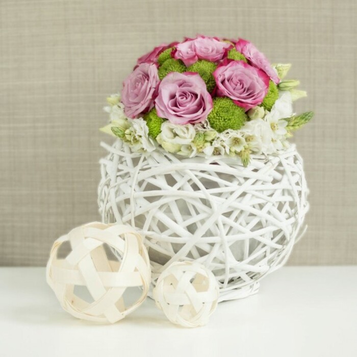 This image showcases an elegant wedding table centerpiece that would sit beautifully among white linens and sparkling glassware in Brent Cross venues. The flowers form a rounded mound, composed primarily of roses in tender shades of pink and lavender, each one fully open so that the petals create soft, layered rosettes. Some blooms exhibit deeper pink edges, adding dimension and drawing the eye gently around the arrangement. Interspersed through the roses are small green button chrysanthemums, their compact, dome-shaped heads bringing a charming texture and a vivid flash of lime-green that feels lively and youthful. At the edges, tiny white filler flowers cluster gently, emerging between petals and foliage to create a feather-light border that softens the bouquet's outline and lends a sense of easy movement. The whole composition is gathered into a distinctive white spherical vase made from intertwined, twig-like strands, giving the impression of a modern rattan basket. Its open lattice structure allows hints of stem and shadow to peek through, and its smooth, painted white surface complements the crisp tabletops often seen in Brent Cross restaurants and event spaces. Just to the left of the main arrangement, two decorative spheres crafted from pale wicker rest on the same white surface, one larger and one smaller. Their open-work patterns echo the weaving of the vase, reinforcing the design theme and adding visual rhythm. The backdrop is a soft blur of warm beige fabric, reminiscent of linen or burlap, which provides subtle texture and a cosy atmosphere without competing for attention. Gentle, even lighting reveals every curve of rose petal and every twist of wicker, giving the centerpiece a serene, polished presence, perfectly suited for wedding receptions, engagement parties, or intimate family gatherings near Brent Cross.