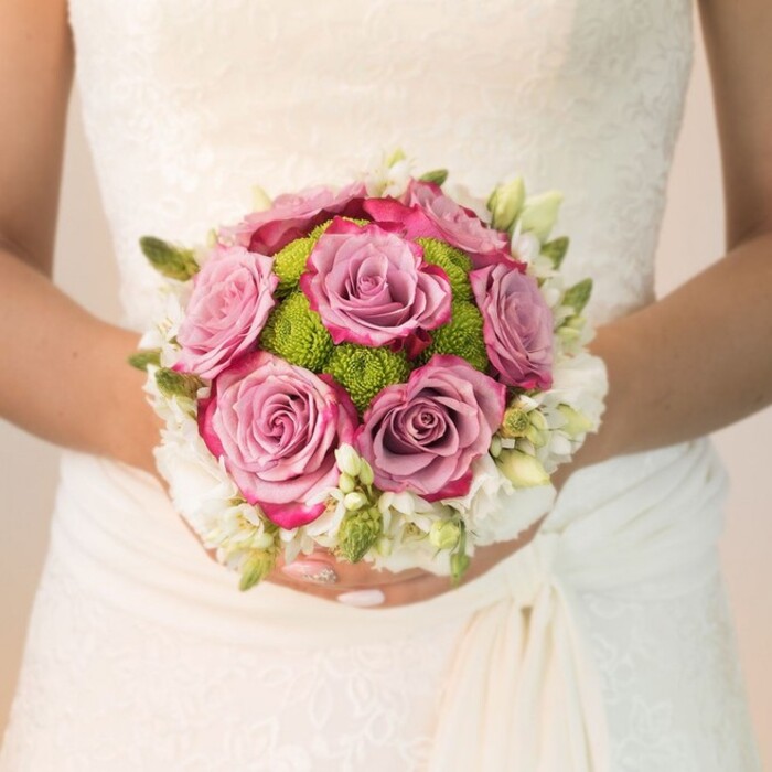 A close-up, natural-light portrait from a Brent Cross wedding frames a bride's hands as she presents her compact, beautifully balanced bouquet against an intricately detailed lace gown. The focus centers on a gathering of dusty mauve and soft lavender-pink roses, each petal edged in subtle fuchsia gradients, reminiscent of the gentle hues that grace Brent Cross at sunset. Nestled beside these classic blooms, radiant lime-green button chrysanthemums offer a lively pop of color and a contemporary flourish, their tight, pom-pom shapes casting playful shadows in the gentle afternoon sun. The bouquet's rounded shape is bordered with delicate clusters of white-very possibly freesia or fine spray roses-enhanced by bright green buds among their stems, lending definition and a refreshing clarity. The gown's lace overlay and draping at the waist evoke a timeless bridal aesthetic, set in modern contrast against the bouquet's textural vibrance. Polished hands, nails painted blush with a hint of sparkle, cradle their floral treasure with care. Assembled by Brent Cross Flowers, this image reflects the joy, beauty, and attention to local detail characteristic of North West London's favourite wedding florists.