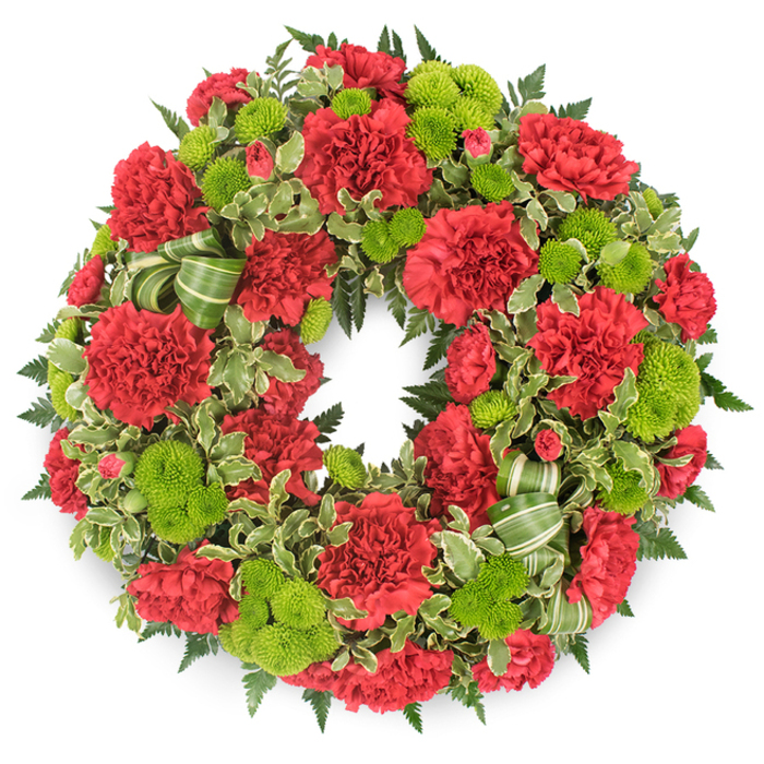 Set against a clean white background, this circular memorial wreath showcases a striking harmony of deep red and bright lime green, crafted with the precision and care of a seasoned florist. The wreath is formed from a dense ring of blooms, with no bare spaces, giving it a full, substantial presence that feels both solid and comforting. Red carnations dominate, their soft, frilled petals layered tightly together to form plush, rounded flower heads. Some are just beginning to unfurl, while others are fully open, their many petals catching the light and creating subtle shadows. The red hue is warm and sincere, an apt choice for a funeral tribute. Threaded among these carnations are clusters of lime green button chrysanthemums, each bloom a small, perfectly rounded cushion of compact petals. The mums add a lively jolt of colour that lifts the design, much like a patch of bright greenery glimpsed amid the busier streets and overpasses near Brent Cross. Supporting the flowers is a bed of mixed foliage: dark fern fronds provide a feathery framework, while broader leaves and sprigs of variegated pittosporum, edged with creamy white, break up the darker greens and introduce intricate patterns. A coordinating ribbon in shades of green, lightly striped, is woven into the lower section of the wreath in fluid loops that echo the circle's continuous form. This wreath would be at home resting beside a coffin during a service, placed at a graveside, or displayed at a memorial gathering in the Brent Cross area. It conveys sympathy, remembrance, and the wish to surround a farewell with living colour and care.