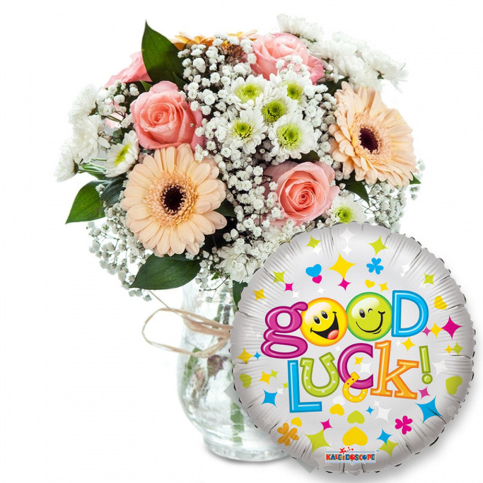 This image presents a joyful, thoughtfully arranged 'Good Luck' gift, photographed on a clean white field so every detail is clear and immediate. Dominating the left and centre is a full bouquet in a clear glass vase, the neck casually tied with a length of natural twine which lends an artisanal touch. The floral palette is soft and uplifting: light pink roses with gently cupped petals provide a romantic core, while peach-toned Gerbera daisies with deep brown centres inject warmth and cheerful formality. Wispy clouds of white gypsophila (baby's breath) drift through the arrangement like lace, and clustered white-and-lime chrysanthemum florets add lively, crisp texture. Deep green foliage frames the blooms, creating contrast and weight. On the right and slightly overlapping the lower vase, a round metallic silver Mylar balloon carries the exuberant Good Luck! slogan in playful, multi-coloured lettering; the two o's are replaced by bright yellow emotive smileys (one winking, one sticking its tongue out), and the surface is sprinkled with colorful stars, hearts and confetti shapes in pink, blue, orange and yellow. A small KALEIDOSCOPE logo is visible at the balloon's base. The lighting is even and natural-feeling, emphasizing the velvety rose petals and the satiny shine of the daisies, while the glass vase picks up soft highlights that enhance clarity. The overall emotion is encouraging and celebratory - an ideal composition for sending supportive wishes before exams, a new job, a move, or any milestone within the Brent Cross neighbourhood and nearby Hendon or Colindale.