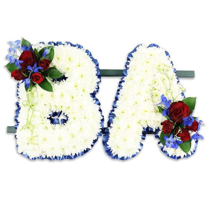 In this solemn, handmade memorial piece the capital letters BA are rendered as two freestanding floral forms, each composed of hundreds of small, immaculate white chrysanthemum blooms that together create a dense, cloud-like surface and a feeling of quiet purity. The edges of the B and the A are traced in a ruffled, royal blue satin ribbon that crisply defines their shapes and lends a restrained, dignified contrast to the white mass. A carefully placed cluster of accent flowers decorates the upper left of the B: velvety deep red roses in varied stages of bloom, punctuated by small, vivid blue delphinium-type florets, and softened by dark green foliage with pale green tendrils that trail gently and lend an organic, natural line. The A is balanced by a corresponding cluster near its lower right, mirroring the reds and blues and tying the composition together visually and emotionally. The pair sits side-by-side on a narrow dark green rectangular stand that keeps the tribute upright and unified, photographed against a neutral white background to keep the focus on colour, texture and craft. Intended for use at a funeral, memorial or commemorative ceremony around Brent Cross, Hendon or Cricklewood, this arrangement speaks of respect and personalised remembrance, marrying traditional sympathy colours with careful floral technique to honour an individual through their initials.
