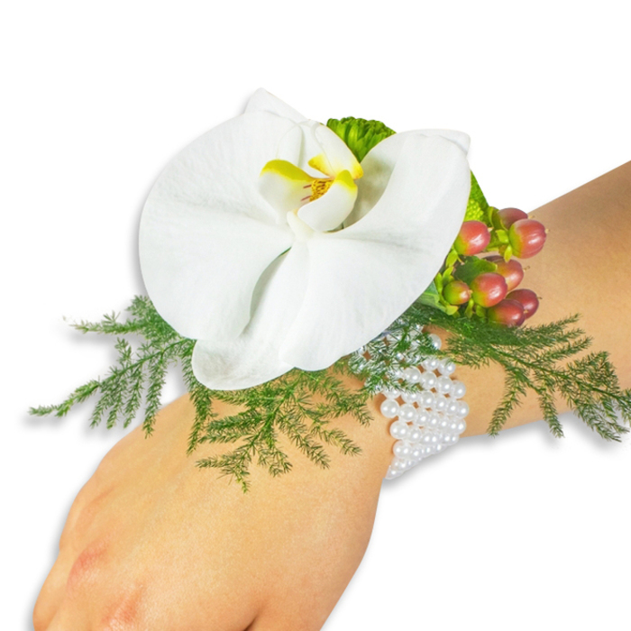 A single white Phalaenopsis orchid composes the centerpiece of a refined wrist corsage, displayed on a pale wrist against a stark white studio background. The orchid's wide, velvety petals fan out like a small sculpture; at its throat a vivid yellow lip, edged with a deeper orange hue, acts as a warm, jewel-like focal point against the pristine white. Nestled beneath and around the blossom, soft asparagus fern fronds create a verdant, feathered cradle that gives the arrangement a pastoral, almost vintage elegance. To the upper right of the orchid sits a compact cluster of Hypericum berries, their skins glossy and richly coloured from pale green to rose and on to a rounded cranberry - a progression that hints at seasonal ripeness and lifts the composition with a quiet pop of colour. A ruffled lime-green accent foliage adds an extra thread of texture beside the berries. The corsage is secured to the wrist with a classic multi-strand bracelet of small white pearls; their subtle iridescence and measured weight lend the piece an heirloom sophistication and feel-good comfort against the skin. The clean lighting and white backdrop draw attention to the interplay of textures - the orchid's satin surface, the fern's lacy filigree, the berries' polished roundness and the pearls' gentle glow - producing a look that is both romantic and composed. Ideal for brides, prom dates or anyone seeking an elegantly restrained accessory, this corsage embodies thoughtful floral workmanship and seasonal freshness. Arranged with local knowledge for the Brent Cross community and ready for delivery across nearby streets and parks, it reads as a small, wearable celebration of craft and quiet beauty.