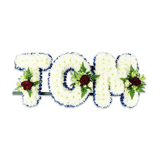 A striking, personalised memorial display spells 'TOM' in bold three-dimensional lettering composed entirely of immaculate white chrysanthemum blooms, each small flower layered tightly to create a cushioned, textural surface that reads as pure and serene. A vivid royal blue ribbon outlines every contour of the letters, its clean line providing striking contrast and lending an air of formality and clarity to the tribute. Within each letter a carefully balanced spray acts as an accent: a single deep red rose anchors the cluster, symbolising enduring love amid loss, while soft ivory spray roses and delicate fern fronds fan around it, introducing nuance of colour and gentle, leafy movement. Visible at the back and base is a narrow dark metal support bar that holds the letters upright for display at a funeral service, chapel or graveside. The photograph places the arrangement against a bright white background under soft, even lighting, which highlights the subtle modulations of petal texture, the sheen of ribbon and the cool green of foliage. This is the kind of custom sympathetic piece often chosen by families in Brent Cross, near Brent Cross Shopping Centre and the nearby residential streets of Cricklewood or Golders Green, when they want a dignified, personal statement of remembrance. Calm, composed and handcrafted with experienced floristry technique, the design conveys reverence and care without excess, offering a heartfelt tribute that reads clearly to mourners and guests alike.