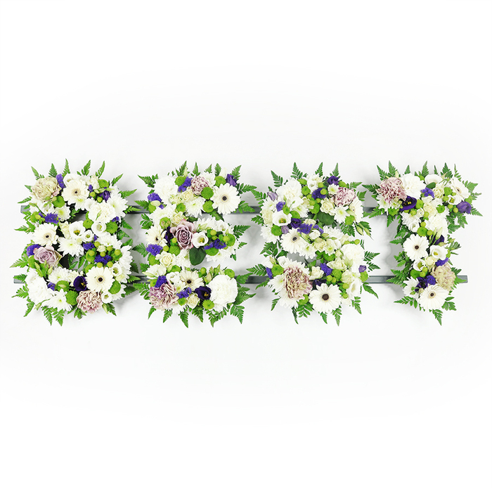 A meticulously composed, horizontal floral display spelling BEST, photographed on a pristine white field so the letters appear crisp and serene. Each capital letter is densely formed from a serene palette of pure whites, soft lavenders, mauves and lively greens. Prominent white gerbera daisies with their pronounced centres sit alongside cloud-like white lisianthus and compact, full white carnations, creating a variety of petal shapes and tactile surfaces. Soft mauve and lavender roses, together with ruffled purple carnations and tiny deeper purple filler blooms, are woven through the composition to add measured warmth and a respectful depth of tone. The edges of each letter are embraced by feathery fern fronds that provide a soft, natural border, while small green button chrysanthemums are nestled into the interior spaces, giving a fresh, grassy puncture of colour and visual weight. A dark grey or black support frame underlies the flowers, subtly visible in narrow gaps and helping each letter hold its sculptural form. The overall effect is one of considered restraint and quiet beauty - a tribute suitable for memorial services and funeral settings where clarity of message and tenderness of execution matter. As a florist familiar with Brent Cross and its surrounding streets, I think of creating pieces like this for families who want a personal, dignified memorial that reads clearly from the front pews and conveys a lasting sense of love and remembrance.