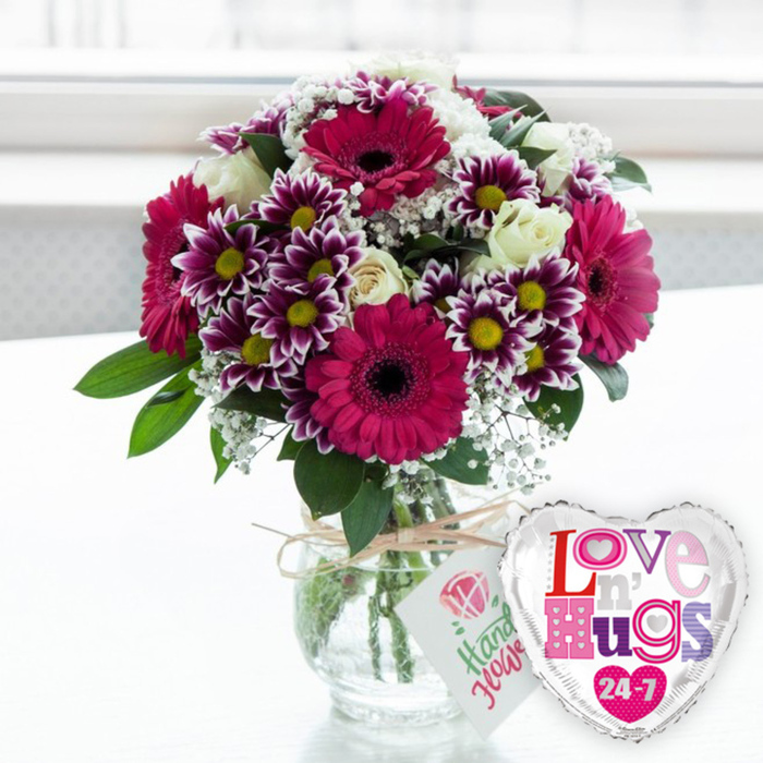 Displayed on a spotless, light-coloured surface that could sit in a living room near Brent Cross station, a vivid bouquet in a glass vase is paired with a heart-shaped foil balloon to create an image of pure celebration. The vase itself is a classic, rounded cylinder of clear glass, filled partway with fresh water. Inside, a tidy bundle of green stems is visible, converging at the base where the florist has grouped them with care. Around the neck of the vase, a simple length of rustic twine has been tied, its natural fibres forming a small bow that supports a rectangular white gift tag. The tag is printed with a minimalist pink rose icon and easy-going green script, a quiet detail that hints at a hand-finished, local design from Brent Cross Flowers. Above the vase, the bouquet swells into a generous, dome-shaped canopy of blooms. Large gerbera daisies in striking fuchsia and magenta shades lead the eye, their smooth, ray-like petals circling dark central disks that give the composition a strong focal point. Threaded between them are two-tone chrysanthemums whose petals transition from saturated purple to crisp white tips, surrounding bright yellow centres like tiny stars. Several creamy white roses are nestled thoughtfully amongst these vibrant flowers, their petals still slightly closed and layered, offering a softer counterpoint and hinting at a subtle fragrance. Sprays of delicate white baby's breath are scattered throughout, creating a gauzy, cloud-like texture, while luscious green foliage peeks from beneath and between the blossoms, completing the rounded outline. To the right of the arrangement stands a glossy heart-shaped balloon edged in reflective silver. Its white interior is filled with fun, multi-coloured letters in pinks, purples and blues spelling out an affectionate Love n' Hugs 24-7, accented with small heart motifs. The softly blurred, bright background suggests natural daylight pouring in, perhaps from a window overlooking the North Circular or nearby Hendon Way, keeping attention firmly on this joyful, love-filled gift ideal for birthdays, anniversaries or just-because deliveries across Brent Cross.