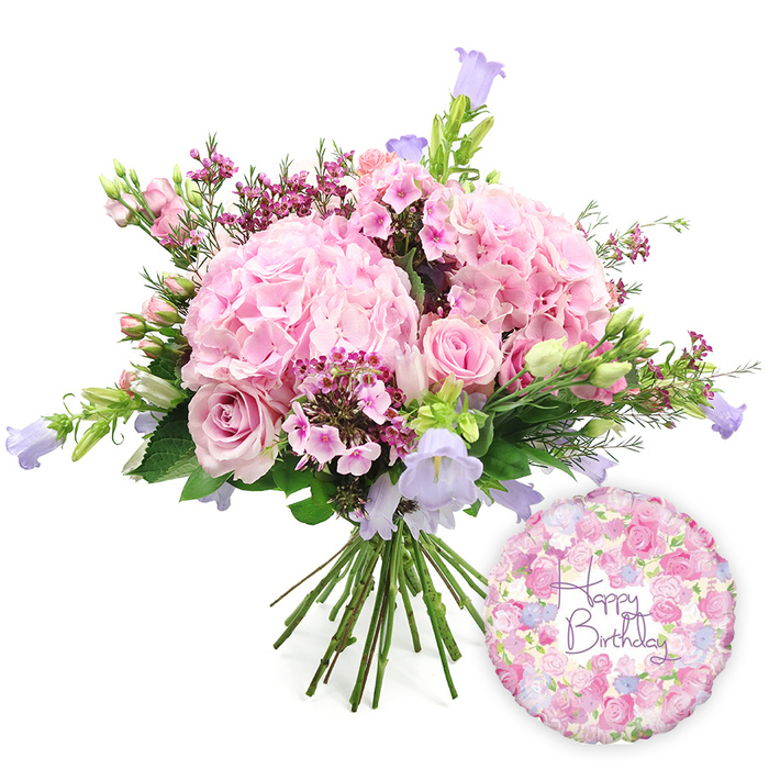 Centred against a bright white backdrop, this photograph showcases a hand-tied birthday bouquet paired with a coordinating foil balloon, creating a complete, celebratory display ideal for marking a special day in Brent Cross. The bouquet itself is lavish and lush, shaped into a classic dome with several substantial heads of blush pink hydrangea providing the main volume. Each hydrangea cluster is formed from countless, closely packed petals that give a velvety, cloud-like appearance, softening the overall silhouette. Interwoven with these hydrangeas are soft pink roses in different stages of bloom: some are wide open, displaying their layered, spiral hearts, while others remain in tighter buds that will continue to open after delivery, bringing ongoing enjoyment. Dotted throughout the bouquet, small sprays of intense fuchsia or magenta waxflowers, or comparable petite filler blooms, introduce tiny points of contrast, depth, and a touch of countryside charm. Slender stems of bell-shaped campanula lift the eye, their lavender and light purple blossoms adding vertical structure and a cool, serene undertone to the predominantly pink palette. Around the flowers, varied foliage provides a verdant frame: slim, pointed leaves mingle with broader, rounded greenery, creating texture and balance that echo the mix of urban and green spaces found around Brent Cross and nearby parks. The stems of the bouquet are clearly visible at the bottom, gathered neatly in a hand-tied fashion, indicating that it is ready to be placed directly into a vase upon arrival. To the lower right, a round metallic foil balloon adds an unmistakable birthday message. Its surface is decorated with a delicate floral pattern of small roses and buds in coordinated pink, cream, and lilac tones, surrounding the elegantly scripted words Happy Birthday in rich purple, completing the joyful ensemble.