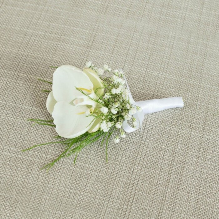 A meticulously crafted white orchid boutonniere is displayed against a softly lit, light beige fabric backdrop with a subtle woven texture, reminiscent of finely dressed tables at an intimate Brent Cross reception. At the centre of the design sits a single Phalaenopsis orchid bloom, its generous, rounded petals pure white and gently curved, like porcelain held in careful hands. The petal surfaces are smooth with a faint sheen, catching the light so that the flower appears almost luminous. In the middle, the orchid's throat reveals a hint of pale butter-yellow, a small accent of warmth that suggests freshness and natural complexity without disturbing the clean, modern palette. Surrounding the orchid are airy sprays of baby's breath, each tiny white blossom forming part of a loose, cloud-like collar that adds lightness and romantic charm, reminiscent of soft mist over nearby parks and gardens. Slender strands of vibrant green foliage emerge from beneath the blooms, fine and featherlike, offering a refreshing contrast and a sense of organic movement, as if a gentle breeze from the streets around Brent Cross station had just brushed past. The stems of the flowers and greenery are drawn together with precision and wrapped in a crisp white satin ribbon, the fabric smooth under the fingers and secured in a small, neat bow, perfectly positioned near the top to keep the boutonniere balanced when pinned to a lapel. The neutral oatmeal background keeps attention on the interplay of white and green, highlighting the design's elegance and simplicity. Crafted with the quiet confidence of a local wedding florist, this piece feels ideal for a groom or groomsman walking into a ceremony in North West London, carrying with him a symbol of purity, commitment, and timeless style.