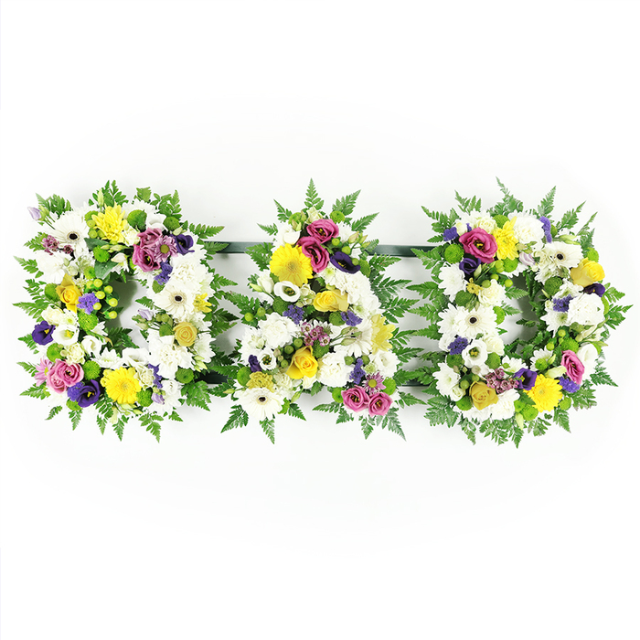 A traditional yet thoughtfully detailed floral tribute spelling the word DAD is shown in full, photographed against a pure white background that keeps the focus firmly on the flowers themselves. The tribute is made up of three separate block capital letters arranged in a straight line, with the two Ds flanking the central A. Each letter is mounted on a hidden frame that is generously edged with lush, deep green fern foliage, the fronds forming a soft, ruffled outline that suggests a natural, garden-inspired style reminiscent of calm corners around Brent Cross. The interiors of the letters are filled to the brim with white chrysanthemums and large white gerbera daisies, packed closely together so that their overlapping petals create a dense, cushiony surface symbolising peace, respect, and remembrance. Breaking up the white are clusters of cheerful yellow gerbera daisies and delicate yellow spray roses or button chrysanthemums, their golden tones bringing brightness and warmth, like filtered sunlight across nearby streets and pathways. Deeper accents of colour come from purple lisianthus and rich magenta or deep pink spray roses, carefully positioned to draw the eye, while tiny lavender filler flowers and bright green button chrysanthemums add detail and gentle contrast throughout the arrangement. All of the blooms are oriented forward so the word DAD can be instantly read from the front, making this a strong focal piece for a funeral, wake, or memorial service. Thin, dark green support bars join the letters along the back, providing structure and alignment without being noticeable from the front. The overall impression is one of a lovingly crafted, bespoke memorial tribute, the sort of arrangement a Brent Cross family might choose to pay their last respects to a beloved father with grace and heartfelt emotion.
