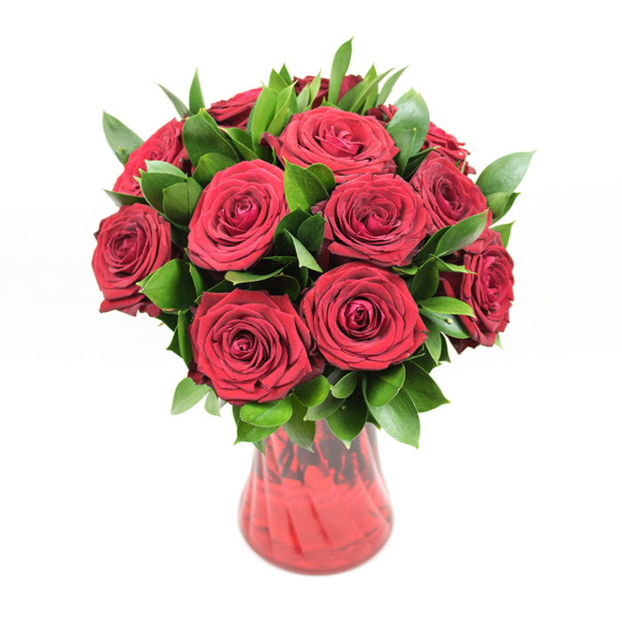 A classic, luxurious display of deep red roses fills a curving red glass vase, shown against a crisp, white background that makes the colours almost glow. Around a dozen to fifteen roses form a compact, rounded bouquet, each flower at the ideal stage of openness: petals unfurled enough to showcase their swirling, velvety centres, yet still tight enough to keep their elegant shape. The red hue is intense and saturated, somewhere between ruby and wine, giving the impression of richness and depth rather than brightness alone. Nestled among the blooms is plentiful dark green foliage, likely ruscus or a similar leaf, with smooth, slightly pointed leaves that catch the light along their glossy surfaces. This greenery breaks up the expanse of red, adds volume and structure, and creates an appealing, natural rhythm throughout the design. The vase itself is made from translucent red glass, its body gently tapering and then slightly flaring at the top, with a subtle ribbed or undulating texture that refracts the light into deeper streaks of crimson. Through the coloured glass, the stems are hinted at rather than sharply visible, adding to the arrangement's sense of cohesion. Lit with soft, even illumination, there are no harsh shadows-only gentle shading that outlines each petal and leaf. The entire image feels intentional, like a professionally crafted studio portrait of a bouquet ready to be delivered for Valentine's Day, a milestone anniversary, or a romantic surprise in Brent Cross or the nearby tree-lined roads towards Golders Green. The mood is unapologetically romantic and expressive, ideal for conveying heartfelt affection, admiration, and the timeless message that only red roses seem able to deliver.