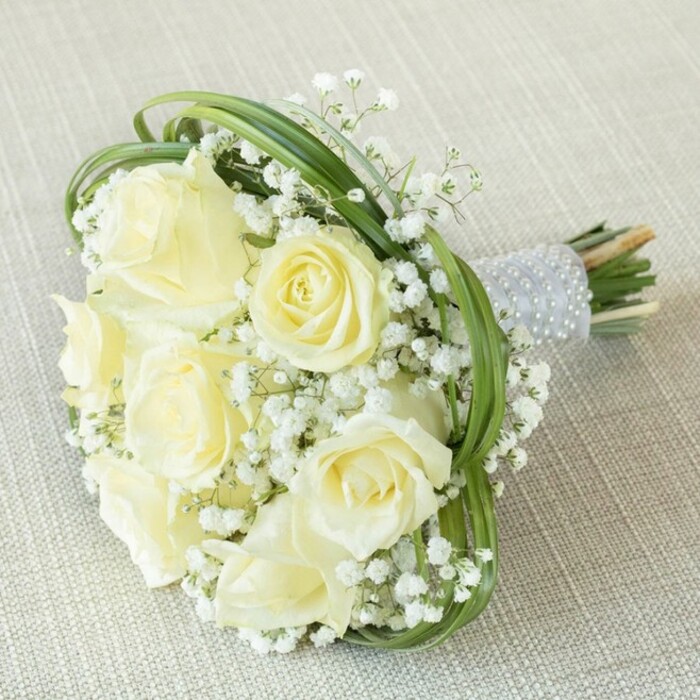 This extraordinary bouquet, displayed against a textured beige fabric reminiscent of the inviting interiors seen in Brent Cross event spaces, is a testament to classic style and local floral artistry. Seven to eight creamy white roses, each at varying stages of blossom, take centre stage-some reserved in tight buds, others lush and radiantly open. These are surrounded by abundant tufts of delicate baby's breath, their miniature white florets providing cloud-like fullness and a subtle contrast in scale and texture. Striking this harmonious balance is a dramatic framing of vivid, arched bear grass, each loop lending a gentle contemporary twist to the tradition-rich arrangement. The stems are bound neatly together, wrapped in sophisticated silver-grey satin ribbon, which is further enhanced with meticulously applied parallel rows of pearly white beads. This touch of luxury is often sought after for weddings and formal gatherings held at Brent Cross's finest venues. Underneath the pearl-strung handle, the cut ends of the green stems peep out, grounding the bouquet in natural beauty. With every detail considered, this arrangement is both a token of purity and a reflection of refined craftsmanship-a quintessential bridal accessory or elegant centrepiece for any meaningful celebration in and around Brent Cross.