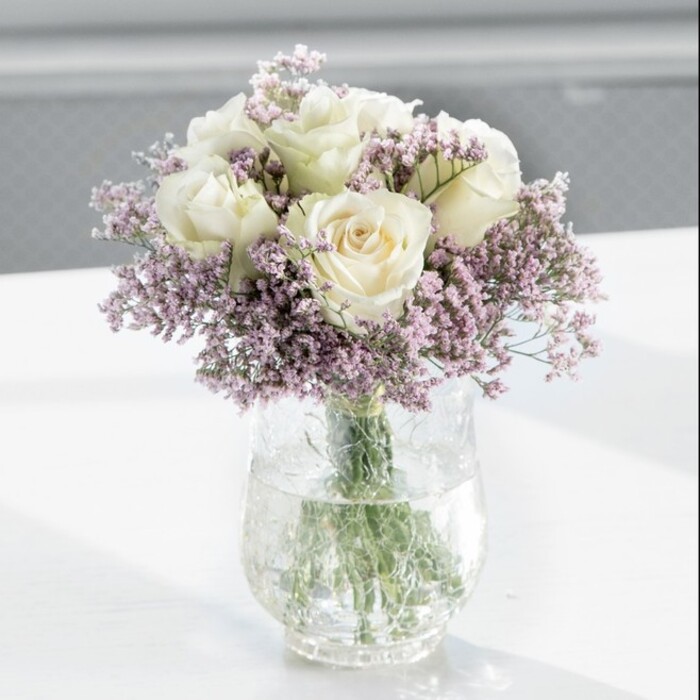 In the centre of the photograph, a lush, round bouquet of creamy white roses and airy lilac statice stands gracefully in a clear glass vase, evoking the quiet elegance of a well-kept Brent Cross townhouse. The roses dominate the composition, roughly a dozen generous blooms gathered closely so their velvety petals almost touch. Each flower is caught just as it begins to open fully, the spiralling layers of petals transitioning from a soft, ivory-cream at the heart to a slightly lighter, almost luminous edge. Tucked between these classic roses are clouds of delicate, lavender-toned statice, their tiny, papery petals appearing in countless clusters. The statice creates an intricate, lace-like veil around the roses, softening the bouquet's silhouette and lending a sense of movement, as though a gentle breeze had brushed through a garden. The arrangement rests in a tall, cylindrical glass vase filled with clear water, which reveals the fresh green stems aligned in a neat bundle. The stems' vertical lines provide a subtle contrast to the rounded dome of blooms above, giving the composition balance and harmony. The vase is positioned on a smooth white surface that reflects slivers of light and the faintest of shadows, enhancing the bouquet's luminous quality. The background is softly blurred, suggesting an airy, daylight-filled room-perhaps a calm corner of a home off Hendon Way or a bright office overlooking Brent Cross Shopping Centre-where natural light washes over the flowers. The overall impression is of serene refinement and heartfelt sentiment, making this bouquet visually suited to occasions like sympathy tributes, housewarming gifts, or meaningful celebrations shared with loved ones in and around Brent Cross.