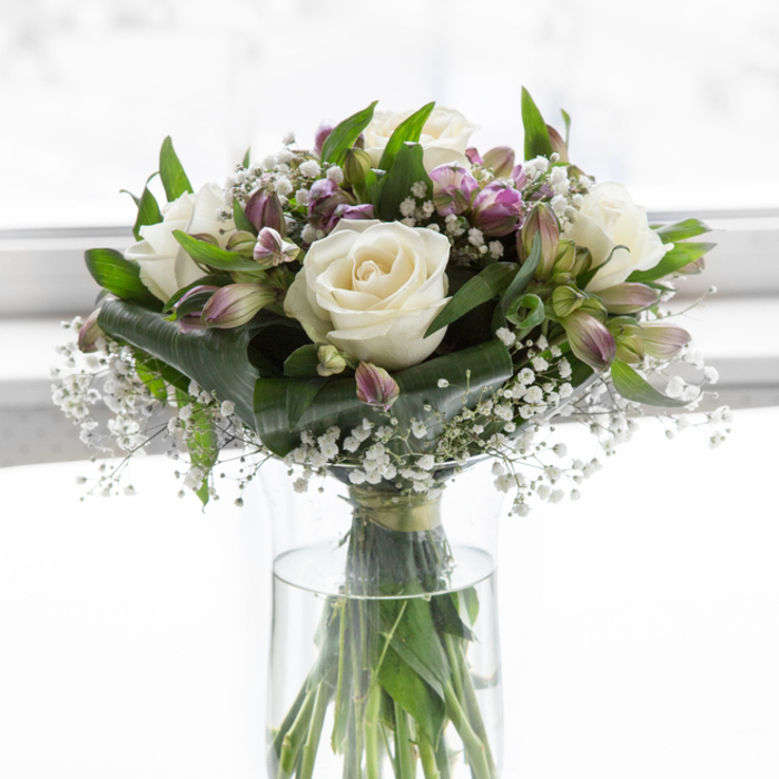 A tall sympathy bouquet in a clear, cylindrical glass vase is shown against a bright, soft-focus white background, allowing every nuance of the flowers to stand out. At the heart of the design, full-headed white roses bloom in calm, measured spirals, each creamy petal curving outward in a way that suggests tenderness and dignity. Between them, alstroemeria in cool shades of mauve, purple, and soft green add subtle layers of colour; some blossoms are open and expressive, while others remain in bud form, hinting at quiet continuity. Fine, frothy clusters of white baby's breath fill the spaces between the larger blooms, their tiny flowers creating a light haze that softens the overall silhouette. Surrounding and supporting the central flowers, rich green foliage provides structure and contrast, from broad, polished leaves that catch the light, to more slender, upright stems that lend the arrangement a gentle vertical line. The clear glass vase reveals the straight green stems immersed in clean water, underscoring the sense of freshness and professional care one might expect from Brent Cross Flowers. The lighting is even and natural, as though the bouquet were placed close to a window in a home off Templehof Avenue or near Clitterhouse Playing Fields, where the sunlight is diffused by sheer curtains. The peaceful palette of whites, greens, and soft purples, combined with the airy composition, creates an atmosphere of calm reassurance. This bouquet feels particularly suited to sending condolences across Brent Cross, placing on a memory table, or offering as a quiet floral presence during a remembrance gathering or reflective moment at home.