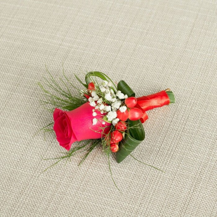 An elegant boutonniere is displayed diagonally across a softly textured, light beige fabric, its gentle weave suggesting the linen often used on wedding tables in Brent Cross venues. The arrangement flows from the lower left to the upper right, drawing the eye first to a striking fuchsia-pink rose at the centre. This rose is in prime condition, with tightly layered petals forming a vivid, velvety rosette that gradually opens outward, the colour deepening in the shadows of the inner spiral. Nestled around the rose are delicate clouds of white baby's breath, their tiny star-like flowers providing a soft contrast that lightens the overall look and adds a touch of classic bridal charm. Among these white clusters, bright red hypericum berries are carefully placed, round and glossy, lending a subtle festive accent that feels perfectly suited to winter weddings or evening receptions near Brent Cross and Golders Green. The foliage plays a vital supporting role: fine, wispy stems of fern-like greenery extend gracefully from the sides, giving a sense of motion, while broader, darker leaves are expertly curled and layered beneath the rose, forming a structured collar that frames the bloom and ensures it will sit neatly on a lapel. The stems are tightly gathered and wrapped in a luxurious red satin ribbon, finished in a neat knot that looks both polished and comfortable against fabric. Soft, even lighting casts gentle shadows that emphasise each contour, highlighting the craftsmanship of a skilled local florist who understands both style and practicality for events across the Brent Cross area, from intimate registry office ceremonies to grand hotel celebrations.