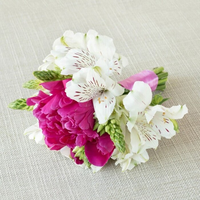 A compact, hand-crafted floral piece lies horizontally on a softly textured, pale beige surface that resembles natural linen or fine burlap, the neutral fabric providing a calm, understated backdrop. At the centre of the arrangement, a lush fuchsia peony bloom opens fully, its petals densely layered and gently ruffled, creating a rounded, cushiony form of rich magenta that instantly commands attention. Around this bold focal flower, several white alstroemeria-Peruvian lilies-unfold in contrast, their slender, slightly flared petals edged in creamy white and brushed inside with slender streaks and speckles of deep maroon, like delicate ink lines. These patterned interiors add intricacy and visual movement, drawing the gaze deeper into the bouquet. Threaded throughout are fresh green buds and narrow, elongated florets, possibly gladiolus or another linear filler, bringing vertical accents and a crisp, botanical texture that keeps the design feeling light and contemporary. The stems are neatly aligned and secured with a bright pink satin ribbon, wrapped firmly but elegantly, its glossy finish catching the light and echoing the vibrant tone of the central peony. The lighting is soft and even, casting gentle shadows that emphasise the velvety depth of the fuchsia petals and the clean brightness of the white lilies, as if the bouquet has just been set down on a dressing table before a wedding in Brent Cross or nearby Golders Green. The overall impression is one of polished romance-an arrangement that could serve as a chic boutonnière, petite bridesmaid posy, or corsage, handcrafted by an experienced local florist who understands how to blend colour, texture, and balance for intimate celebrations across north-west London.