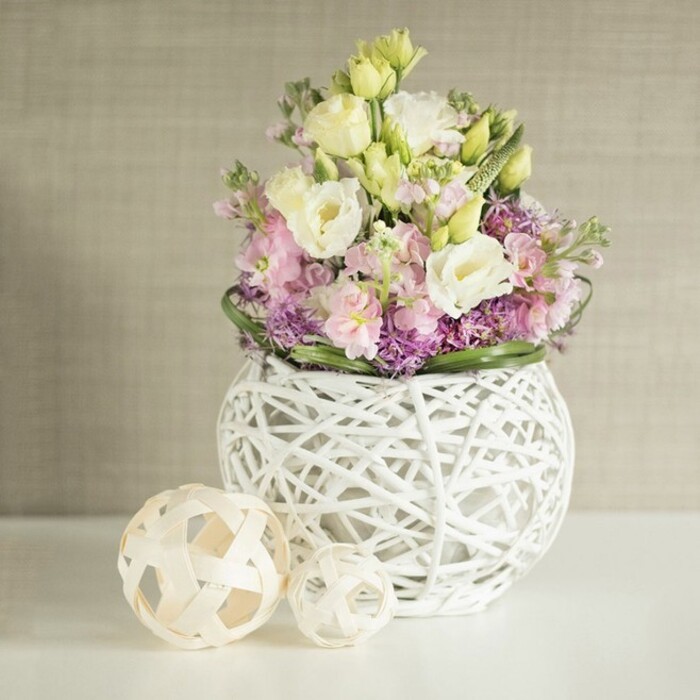 This refined table arrangement, a true signature of Brent Cross floral artistry, features a lush medley of pastel blooms cradled within a modern, airy white vase-its construction reminiscent of intricately woven rattan or slender painted twigs. At the bouquet's core, ruffled ivory and soft yellow eustoma unfurl amid elegant pink matthiola, their stately stems rising to bring vertical grace and a touch of romance reminiscent of spring markets off Tilling Road. The lower layers burst with clusters of small, vibrant purple blooms-likely asters or decorative alliums-that ground the display, while a circlet of fresh, deep green foliage lends a sense of fullness and vitality. To the left, resting on the pristine white table, two decorative spheres echo the vase's woven design in pale natural rattan, elegantly reinforcing the room's handmade, artisanal ambiance. All is set against a warm beige backdrop whose gentle vertical texture suggests high-quality linen wallpaper, infusing the space with both modernity and timeless comfort. Gentle, diffuse light flows through the scene, heightening every soft curve and pastel hue. This arrangement is ideal inspiration for spring wedding tables in Brent Cross or sophisticated home gatherings, blending locally sourced freshness, modern design, and the inviting atmosphere of a community that treasures elegant yet natural interiors.