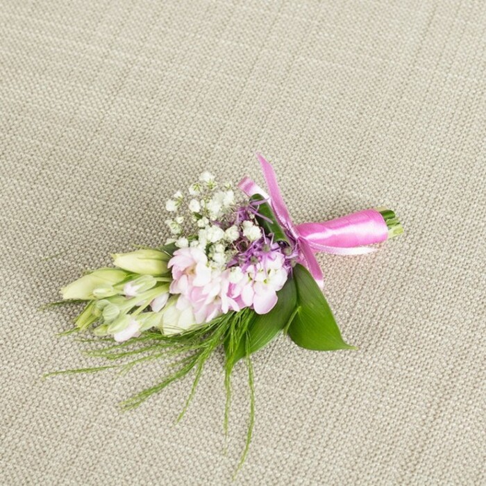 A lovingly arranged boutonniere or small corsage is displayed against a subtly textured, light beige linen backdrop, the fabric's fine weave creating a calm, neutral canvas that allows every colour and shape to be clearly sensed. At the centre of the design, you can imagine a handful of soft, blush-pink blossoms, similar to miniature stocks or ruffled garden flowers, their many petals gathered tightly into rounded clusters. These petals carry a gentle ombré of pink, deepening slightly as they fold inward, suggesting softness and a freshly opened bloom. Nestled among them, tiny white baby's breath florets scatter like delicate stars, forming a light, airy veil that contrasts beautifully with the fuller pink heads. Here and there, small accents of lavender or muted purple flowers appear, their fine, spiky forms adding a subtle depth of colour, much like the shaded corners of nearby Brent Cross gardens in late afternoon. To one side, a small spray of elongated buds, creamy white tipped with pale green, stretches outward; they resemble freesias or similar slender buds, closed but plump with the promise of fragrance. The flowers are supported by broad, glossy green leaves that cradle the arrangement, their smooth surfaces and rich emerald colour grounding the soft pastels. Wisps of fine, hair-like greenery and fern-textured foliage extend beyond the main cluster, lending a natural, slightly wild edge as though freshly gathered from a quiet corner near Brent Cross shopping and residential streets. At the base, the stems are bundled with precision and wrapped in a bright fuchsia satin ribbon, tied into an elegant, symmetrical bow whose sheen suggests both care and celebration. The overall composition feels serene yet joyful, a handcrafted detail perfectly suited to weddings, proms, or formal events where guests might be receiving flowers delivered locally by Brent Cross Flowers.