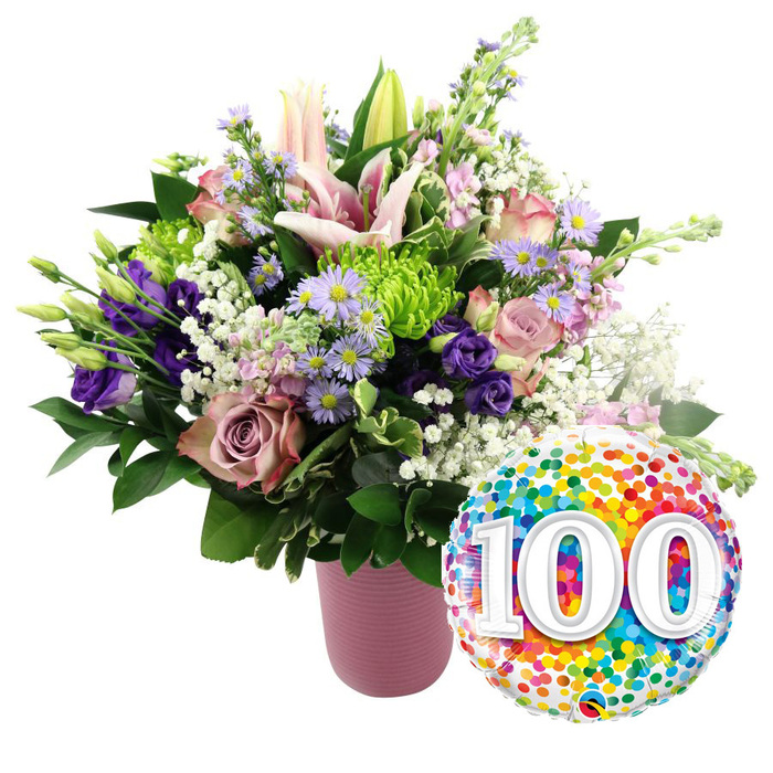 This image captures a radiant centenary tribute: a lavish bouquet in soft, feminine tones paired with a cheerful milestone balloon, all set against a crisp white background. The flowers sit in a modern pink vase, cylindrical in shape with a gentle ribbed texture that catches light in shallow ridges. Its dusty rose hue offers a calm, elegant base for the vibrant arrangement above. Rising at the centre are pale pink Oriental lilies, their elongated buds reaching upwards while several blooms have already opened wide, revealing star-shaped petals with subtle inner shading and long, arching stamens tipped with pollen. Nestled closely around the lilies are roses in complementary shades of blush and mauve, their velvety petals unfolding in generous, romantic spirals. Deep purple lisianthus flowers weave among the roses, adding a richer tone and softly ruffled edges that create movement and interest. Sprinkled throughout the bouquet, clusters of tiny lavender asters with sunshine-yellow centres bring a touch of countryside charm, as if gathered from garden borders near Brent Cross. Bright lime green button chrysanthemums, tightly layered and perfectly round, dot the arrangement and lend a playful, modern twist. Wisps of white baby's breath float between the larger blooms, softening the transitions and adding lightness, while dense, dark green foliage anchors the design, giving it a full, balanced silhouette. To the lower right of the bouquet, a round foil balloon gleams, its surface entirely covered with bright confetti dots in red, orange, yellow, green, blue, and purple. At its centre, the number '100' is written in large, bold white numerals with a faint 3D shadow, instantly conveying the importance of the occasion. Crafted with the care of an experienced Brent Cross Flowers florist, this combination feels perfect for celebrating a 100th birthday or anniversary with family gathered locally.