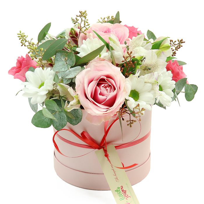 Set against a pristine white background, a pastel pink round hatbox brims with a carefully styled bouquet, capturing the essence of modern floral design that feels right at home in Brent Cross's mix of urban living and green spaces. The hatbox is cylindrical, its matte blush surface smooth and understated, acting as a soft base for the flowers above. A narrow satin ribbon in coral-red wraps around the middle and is tied into a tidy bow at the front, lending a neat finishing touch. From the knot of the bow, a cream gift tag hangs lightly, decorated with green script in a handwritten style that hints at a personal message or the signature of Brent Cross Flowers. The blooms themselves form a lush, dome-shaped canopy over the box's edge. Several large, light pink roses, some with deeper fuchsia tones at their centres, create a romantic focal point; their petals are layered and open, giving the impression of fullness and gentle movement. Surrounding these are clusters of white daisy-like chrysanthemums, each with a bright yellow-green centre that adds a sense of brightness and innocence. Among the main blooms, slender white alstroemeria with subtly marked inner petals weave through, their trumpet shapes providing elegance and airiness. Deeper, vibrant pink flowers - most likely lisianthus or small spray roses - punctuate the arrangement with rich accents of colour. The bouquet is interlaced with greenery: distinctive silvery eucalyptus leaves arc and overlap, mingling with slimmer, darker green foliage and a few reddish-brown buds or seed pods that introduce depth and a slightly wild, natural texture. Altogether, the composition feels fresh, balanced, and inviting, ideal for gifting on birthdays, anniversaries, or Mother's Day, or for brightening a home near Brent Cross Shopping Centre or one of the quieter streets leading towards Hendon and Golders Green.