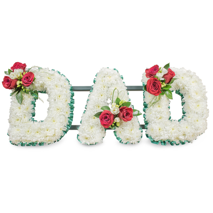 A classic, handmade memorial arrangement that reads DAD in large, freestanding letters, each formed from a dense mass of immaculate white chrysanthemums so closely set that the surface appears seamless and softly sculpted. The chrysanthemums' layered petals create a serene, cushion-like texture, their snow-white tone catching soft studio light against a spotless white background. Every letter is edged in a pleated, emerald-green ribbon that crisply defines the block shapes and echoes the fresh greenery tucked into the posies. Atop the first D sits a thoughtful cluster of deep red roses in varying bloom stages paired with petite white freesia and rich green foliage, offering a focal point that draws the eye and adds warm colour. The central A bears a matching spray near its lower left stem with a slender sweep of green leaves, while the final D is balanced by a similar red-and-white cluster on its upper curve, creating a harmonious, mirrored composition. Narrow, dark green or black metal bars are discreetly positioned behind the letters to hold them together and allow safe placement at a funeral chapel or graveside. The overall impression is hushed and respectful-white chrysanthemums symbolising devoted love, red roses conveying deep affection-crafted with the calm care you would expect from a local Brent Cross florist. At Brent Cross Flowers we prepare tributes like this with sensitivity for families in the area, whether the service is held near the shopping centre or at a cemetery towards the Welsh Harp.