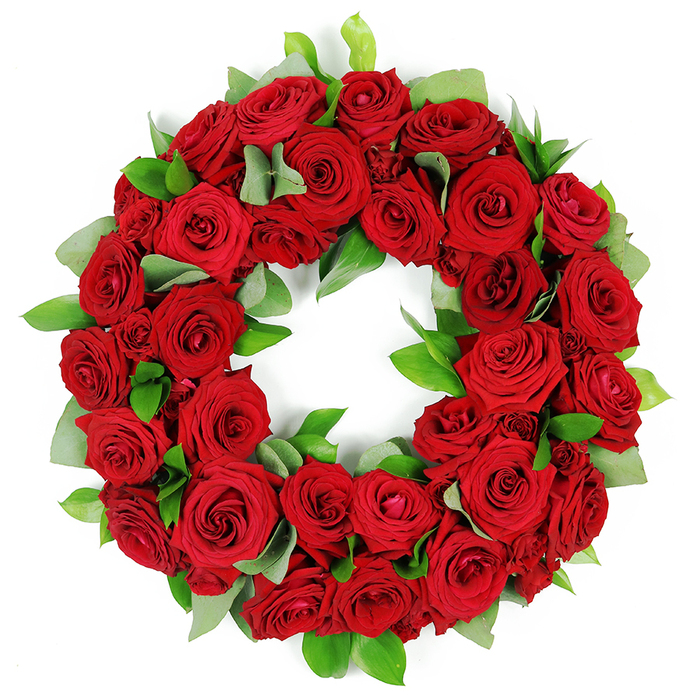 An elegant circular wreath, built from dozens of vibrant, deep red roses, rests on a crisp white background, its blooms densely packed to create a lush, three-dimensional ring. The roses unfold from tightly furled buds to open, velvety petals, each with a saturated hue that seems to glow from within as light skims the edges. Interspersed among the red blooms, clusters of fresh green foliage provide contrast: broad, matte eucalyptus-like leaves breathe against the glossy, pointed ruscus, giving the arrangement structure and depth. The careful layering makes the wreath feel both substantial and refined, a piece that can anchor a memorial display or accompany a funeral setting with quiet dignity. In the context of memorial design, the symbolism of red-love, respect, remembrance-plays against the natural green of leaves, suggesting renewal and continuity. This wreath's presence on a white backdrop makes its colors sing and ensures every petal's texture, from the gentle curves to the delicate veining, is discernible. It is a timeless choice for ceremonies across Brent Cross, Cricklewood, and Golders Green, whether marking a life's passing or honoring a lasting legacy. Crafted with care by a local florist, it speaks to the community's values and the artistry of floral tribute, from the first moment a viewer notices the ring to the last, lingering impression of the deep red petals against pristine white.