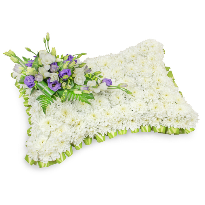 A deeply considered sympathy cushion, shaped into a rectangular pillow and lovingly composed of hundreds of immaculate white chrysanthemum blooms, occupies the frame against a pristine white background. The chrysanthemums form a dense, even carpet of tiny petals that reads visually as softness itself - a soothing plane that suggests rest and reverence. Around the perimeter a soft, light-green satin ribbon is carefully pleated into a scalloped trim, the pale verdant tone offering a subtle, refreshing contrast to the sea of white and neatly defining the arrangement's silhouette. Nestled across the upper-left corner is a discrete but expressive spray of accent flowers: layered white lisianthus sit beside striking purple lisianthus and several still-closed buds, their cooler mauve-purple notes adding depth and a slow, graceful poignancy. Sprigs of delicate fern and broader glossy foliage are entwined through the cluster, lending a lively texture and a sense of naturalness; a darker green leaf wrap secures the bouquet's base with quiet dignity. Soft lighting casts a faint shadow beneath the pillow, emphasizing its three-dimensional puff and the florist's meticulous shaping. When held near, the tribute has a muted floral scent - faint chrysanthemum sweetness balanced with green, leafy freshness - the kind of quiet fragrance that suits a chapel or graveside setting. This arrangement is suited to intimate memorials and formal services alike and is a familiar, respectful choice for families arranging tributes across Brent Cross and the nearby communities of Cricklewood and Staples Corner.