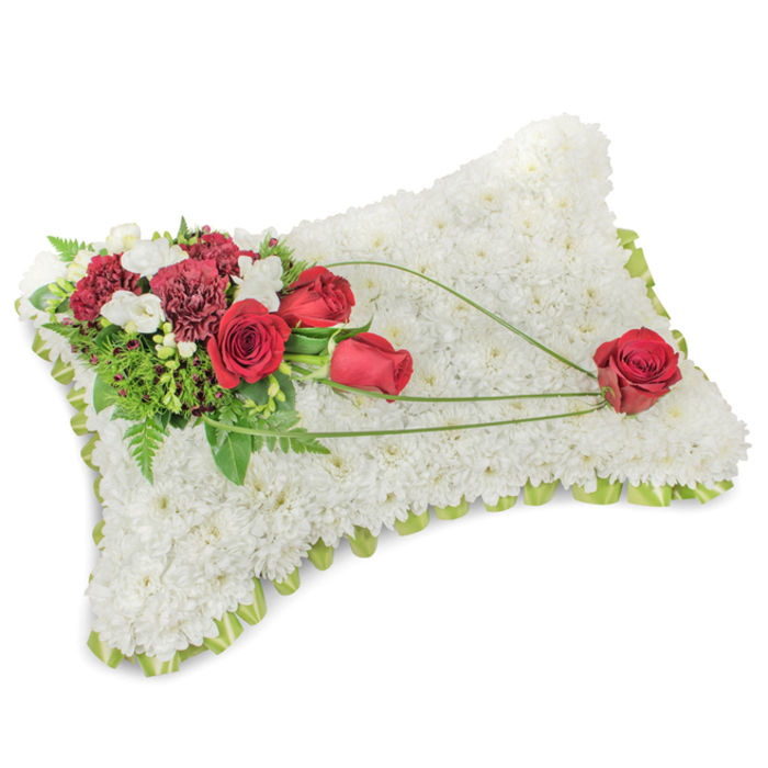 A serene rectangular pillow fashioned from a hundredfold of small white chrysanthemum heads rests in clean, soft light, its textured surface resembling a blanket of snow or fine wool. The chrysanthemums are tightly arranged to form a plush, continuous canvas of pure white, a visual metaphor for peace and comfort. Along the pillow's rim a subtle line of pale green ribbon or foliage peeks through, providing a delicate frame that gently cushions the composition. At the upper left a vibrant floral cluster brings dramatic colour: several deep red roses-some newly opening, others fully unfurled-stand with dark burgundy carnations and tiny white spray blooms that could be freesias, offering both scent and refined contrast. The foliage here is abundant and varied, from airy fern fronds to more substantial glossy leaves, creating dark green recesses that make the red tones sing. From that top grouping, three slender, arching blades of bright green bear-grass sweep diagonally across the white chrysanthemum field toward the lower right corner. These graceful arcs lead the eye and impart a sense of movement and connection, as though the arrangement is carrying a message across the pillow. A single, carefully selected red rose rests at the trail's terminus, poised and emblematic of enduring affection and remembrance. Viewed in subdued natural light-such as the quiet of a nearby chapel or a funeral parlour in the Brent Cross area-the arrangement reads as a timeless and respectful memorial cushion, chosen to express sympathy, honour and lasting love for the person being remembered.