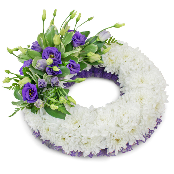 A refined, circular funeral wreath shown against a pure white studio surface, the design composed to convey dignity and calm. The foundation is a dense, unbroken ring of snowy white chrysanthemums; their layered petals form a soft, cushion-like field that reads as both tactile and tranquil. Crowning the wreath's upper-left quadrant is a lush accent of purple lisianthus (Eustoma) in a range of tones from deep violet to pale lavender, with open blooms revealing their layered centres and adjacent buds providing a sense of unfolding life. Interwoven with the purple flowers are sprigs of fresh greenery - some glossy, variegated leaves and some lighter, feathery fern fronds - which introduce texture, cool colour contrast, and a natural rhythm to the composition. A narrow, crinkled ribbon or mesh in dark purple peeks from under the main body along the inner and outer edges, echoing the lisianthus hue and adding a subtle, refined border that frames the white chrysanthemum ring. The photographer used soft, diffuse lighting that casts a gentle shadow beneath the wreath, enhancing its three-dimensional form and the layered quality of the blooms. Sensory details suggest a faint floral scent, the clean sweetness of chrysanthemums tempered by the velvety, almost herbal note of lisianthus, creating a calming presence suitable for a memorial setting. This wreath has been assembled with careful attention to balance and proportion to serve as a dignified tribute at a funeral or memorial service. Available for delivery across Brent Cross and neighbouring Golders Green and Mill Hill, it is intended to offer quiet comfort and lasting remembrance to those who mourn.