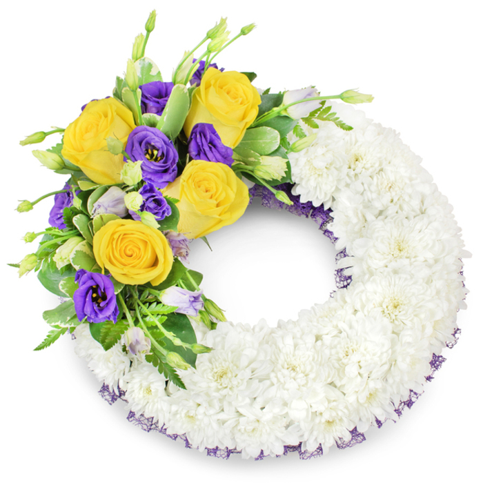 A carefully composed circular funeral wreath, shown against a stark white background, whose majority consists of tightly clustered white spray chrysanthemums forming a smooth, plush ring reminiscent of a pale cloud. The flowers are arranged to create a continuous, reverent surface with depth and a subtle satin sheen that catches ambient light. At the upper left quadrant a pronounced accent cluster arcs gracefully toward the top: several fully opened yellow roses, glowing in soft butter and golden tones, are contrasted by rich, velvety purple lisianthus with their distinctive ruffled cups. Unopened lisianthus buds in paler lavender and touches of green add an organic rhythm, while varied foliage - glossy rounded leaves, delicate fern fronds and faintly variegated greens - provides structure and cooling contrast. A narrow trim of deep purple mesh peeks from beneath the chrysanthemum base along both inner and outer edges, quietly repeating the purple notes and lending a subtle handcrafted finish. The wreath's open centre preserves its traditional form and invites a contemplative focal point. To those arranging memorials in the Brent Cross area - from local parks to nearby community services - this design reads as both respectful and consoling: white for peace, yellow for remembrance, purple for dignity, all combined in a composition that speaks of careful floristry and heartfelt farewell.