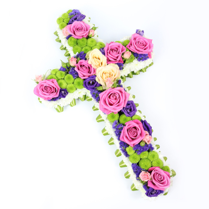 A meticulously crafted floral cross stretches diagonally across a brilliant white background, the slight tilt giving a sense of depth and movement, as though it has just been set down in a tranquil chapel. The cross is framed by a deep border of white spray chrysanthemums, hundreds of tiny blooms packed tightly together to form a soft, continuous cushion that clearly outlines the traditional Christian shape. Just beyond the white fringe, a ribbon of pale-green material-either satin ribbon or carefully folded foliage-wraps around the edges, its subtle sheen adding polish and a delicate whisper of colour, echoing the greenery of parks and gardens around Brent Cross. Inside this crisp frame, the interior of the cross becomes a vivid patchwork of flowers. Large roses in hot fuchsia pink and gentle creamy yellow stand fully open, their layered petals forming generous rosettes that serve as focal points along the arms and down the vertical stem. Bright lime-green button chrysanthemums punctuate the design in neat, rounded clusters, their pom-pom shape providing playful texture and lifting the arrangement with a burst of freshness. Threaded between and around these bolder blooms are clusters of smaller purple and deep violet flowers, most likely lisianthus or statice, which weave through the spaces and add a rich, jewel-toned depth. The lighting is bright yet soft, ensuring every detail of the floristry-each petal curve, each green fold, every compact chrysanthemum head-is clearly defined. The overall impression is of a tribute that honours faith and memory while also celebrating the individuality of the person remembered, a cross suited to funerals, memorial services, or graveside remembrances for families in the Brent Cross area who wish to express both sorrow and enduring love through flowers.