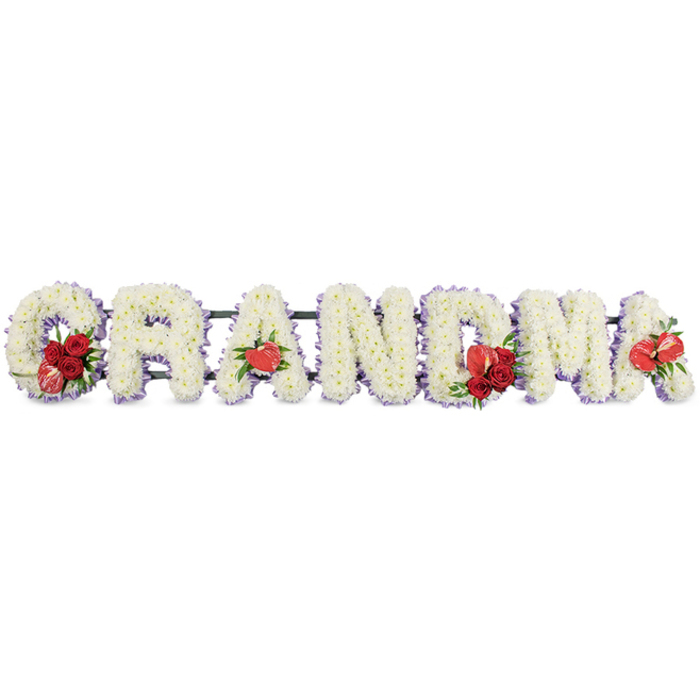 This detailed floral tribute spells out the word GRANDMA in full, each letter rendered in generous, three-dimensional block form and arranged in a straight horizontal line. The letters are completely covered in snowy white chrysanthemums, their compact, cushion-shaped blooms meticulously layered so that every surface is plush and even, creating a sense of softness that feels tender and comforting. Around the outside of each letter, a narrow trim of soft lilac or pale purple ribbon appears just at the edges, subtly outlining the shapes and lending a delicate hint of colour that suggests calm, spiritual reflection. The overall impression is of purity and serenity, yet the tribute is gently enlivened by bold red accents. On the left, the G is adorned with a cluster of velvety deep red roses and a distinctive red-and-white calla lily nestled into its curve, the contrast between the white petals and the rich reds evoking deep affection and cherished memories. The first A features a single, glossy red anthurium at its base, its heart-like form and smooth, waxy texture drawing the eye. In the middle, the M is highlighted by another spray of red roses accompanied by a red anthurium, before the final A repeats the motif with one last anthurium bloom, bringing a sense of rhythm and continuity to the design. Supported by a low dark green frame that holds the letters steady while remaining unobtrusive, this GRANDMA tribute reflects the kind of bespoke funeral arrangement families across Brent Cross, Cricklewood, and Finchley often choose to honour a beloved grandmother, offering a quiet yet deeply personal statement of love at a funeral or graveside service.