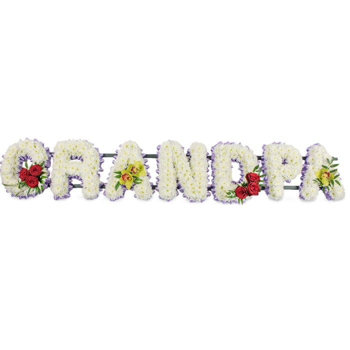 A striking sympathy arrangement spells out GRANDPA in bold, block-style letters, placed horizontally so the word stretches evenly from left to right against a stark white background. Every letter is packed with small white chrysanthemums, arranged so meticulously that the flowers form a smooth, continuous surface without visible gaps. This dense carpet of blooms gives the tribute an inviting, cushioned appearance, conveying softness, purity, and the calm of quiet reflection. Along the edges of each letter runs a thin ribbon of pale lavender or light purple, providing a gentle outline that sharpens the readability of the word and adds a hint of subtle colour, not unlike the soft tones seen in evening skies over Brent Cross. Within this expanse of white, carefully positioned accents add warmth and meaning. The sweeping curve of the initial G holds a compact cluster of deep red roses, surrounded by glossy greenery that sets off their velvety petals. The inner curve of the D mirrors this design with another group of rich red roses, visually linking the beginning and centre of the word. The third letter, the first A, and the final A each showcase a spray of yellow-green orchids, some touched with faint reddish markings, bringing an elegant and slightly exotic note that suggests individuality and special memories. A discreet dark framework and connecting bars sit behind the flowers, holding the letters firmly together as one complete tribute, ready to be displayed at a funeral service, wake, or graveside committal in and around Brent Cross. Overall, the piece radiates dignity and enduring affection for a much-loved grandfather, combining white for peace, red for love, and orchid tones for lasting honour.
