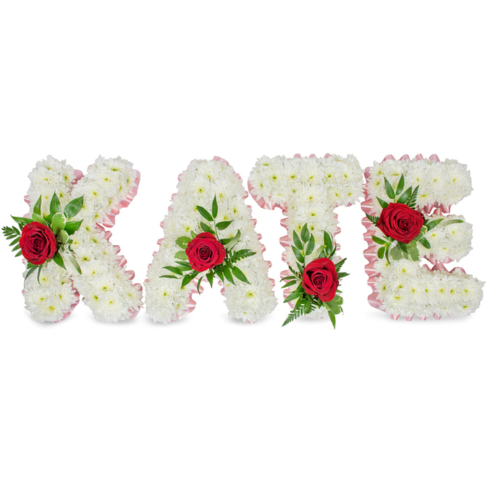 A solemn yet tender floral lettering tribute reads KATE, each capital letter hand-formed from a thick, immaculate mass of white chrysanthemums that produce a soft, pillowy surface and a feeling of calm. Around the edges of every letter a pale pink satin ribbon is carefully pleated and applied, giving a gentle blush to the silhouette and a refined finish under soft, even lighting. Centrally placed on the face of each letter sits a single deep-red rose - vivid, velvety and richly coloured - which becomes the eye's anchor against the white chrysanthemum background. The roses are cushioned by an assortment of fresh green foliage, including broad, glossy leaves and delicate fern-like sprigs that add texture and a sense of life. Placement is intentional: the K's rose rests on the lower left vertical stroke, the A's bloom crowns its triangular peak, the T's rose is set at the base of the left side of its crossbar, and the E's flower is embedded on its middle horizontal bar. The overall palette - pure white, warm pale pink trim, vibrant crimson and deep green - speaks quietly of love, purity and remembrance. Having arranged many tributes for families in Brent Cross, Dollis Hill and around the shopping centre, I picture this piece standing by a casket or on a remembrance table, its refined craftsmanship offering comfort and a deeply personal farewell.