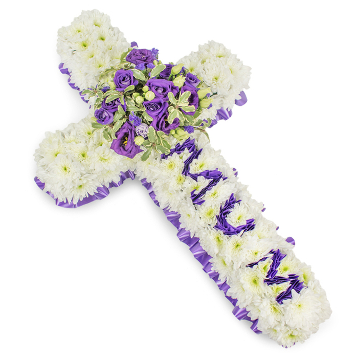 A large, carefully composed Christian cross fashioned from tightly packed white chrysanthemums, photographed against a stark white backdrop so the construction's details are crisp and readable. The chrysanthemums form a seamless, cushiony surface-petals layered to achieve fullness and a soft, consoling silhouette. Around the whole cross a bold, pleated purple satin ribbon has been worked in as a border: its ruffled edge moves from soft lavender to deeper purple, lending a quiet ceremonial trim. On the vertical arm, the name MUM is precisely formed from small purple flower heads-likely lisianthus or miniature spray chrysanthemums-each letter shaped with clear spacing to be immediately legible. Anchoring the design at the intersection, a lush central spray serves as the focal point: intense violet roses with compact buds, lighter mauve lisianthus with delicate, ruffled edges, variegated pittosporum-like foliage with pale margins, and small green berries woven through for texture. Together these elements build a layered visual depth-cool purples nestling into pristine whites while the foliage offers a fresh, living counterpoint. The bouquet suggests a soft, restrained fragrance and a mood of reverent reflection appropriate for funeral services, memorial receptions or graveside tributes. The craftsmanship shows an eye for both aesthetic balance and durability, so the tribute sits respectfully on display in chapels or on cemetery plinths. Locally this style is suited to families arranging services near Brent Cross Shopping Centre, Hendon Park and the quiet lanes of Childs Hill, where such a cross can carry a personal message of loss and lasting affection with understated elegance.
