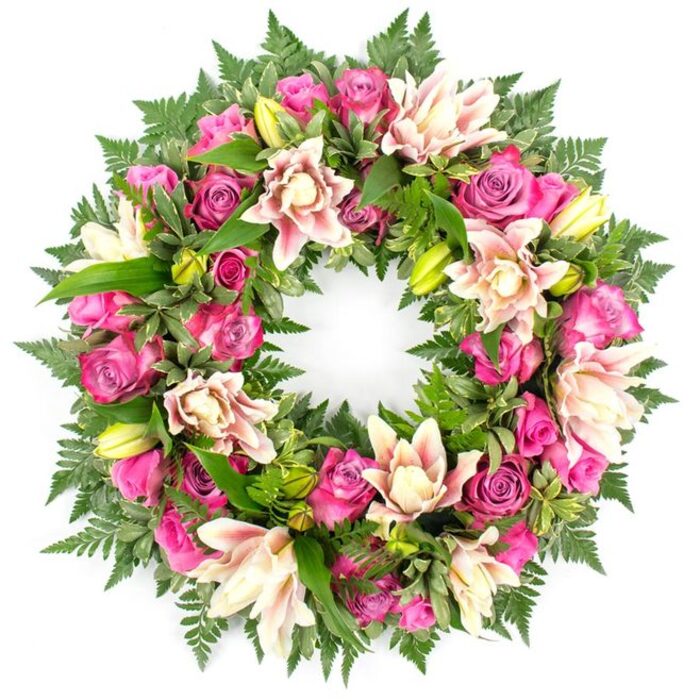 A beautifully composed circular funeral wreath stands out sharply against a spotless white background, its rich colours and textures carefully balanced to convey both sorrow and affection. The wreath forms a flawless ring, fully covered in blossoms and foliage. Dominating the arrangement are roses in an array of pink shades, from intense magenta and bright fuchsia to more subdued, romantic blush tones. Many of the roses are fully open, their layered petals forming plush rosettes that invite the eye to linger; others are semi-open or still in bud, which adds gentle variety and a sense of natural movement. Intermingled with the roses are large, double-petalled lilies, whose broad, ruffled petals overlap one another in soft waves. These lilies are coloured in creamy white, pale pink, and light peach, with occasional deeper pink accents that look like brushstrokes along the edges. Scattered throughout the ring, lime-green lily buds emerge, their sleek, tapering shapes bringing freshness and a subtle upright rhythm to the composition. The entire wreath is anchored by a generous foundation of dark green foliage. Fine, feathery fern fronds frame the outer edge, their lacy texture creating a soft halo, while glossy broad leaves, some with slight variegation, fill the inner spaces, making the wreath appear full and rounded from every angle. Against the clean white backdrop, the combination of deep green foliage and warm pink and cream flowers appears vivid and almost luminous, as though the wreath had just been finished in the studio at Brent Cross Flowers before being delivered to a service near Hendon Way or the Brent Cross shopping centre. The piece radiates care, craftsmanship, and a quiet sense of reverence, making it a suitable and comforting floral tribute for funerals, wakes, and remembrance ceremonies throughout the NW11 and NW4 areas.
