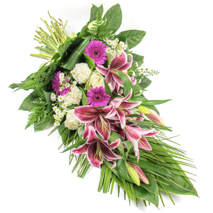 An elegant funeral sheaf, photographed against a clean white backdrop, stretches diagonally from the upper left to the lower right, creating a flowing crescent shape that feels both gentle and composed, like a floral embrace offered to families in Brent Cross. The focal blooms are bold Stargazer lilies, their star-shaped flowers painted with intense fuchsia-pink centres and framed by bright white edges, each petal sprinkled with deep burgundy speckles. Some lilies are open and dramatic, while others remain in slender green buds brushed with hints of pink, suggesting memories still unfolding. Surrounding the lilies, vibrant magenta gerbera daisies introduce a contemporary touch, their tidy petals and cheerful faces adding a sense of fond remembrance. Creamy white roses nestle among them, appearing in various stages from tightly closed buds to delicately opening blooms, symbolising purity, love, and profound respect. Soft white carnations with frilled petals fill the spaces between the larger flowers, lending a cushion-like texture that feels gentle and consoling. Scattered throughout are small clusters of white daisy-like filler flowers, adding a fine, lacy detail and a sense of innocence and light. The arrangement is anchored by an abundance of greenery: broad, glossy dark leaves provide structure and depth, while feathery ferns break up the contours and add movement. Long, slender leaves sweep outward and cascade towards the lower right, emphasising the spray's elongated, teardrop silhouette. At the top left, the bound stems are clearly visible, a reminder of the careful hand-tied work carried out by an experienced florist before the tribute is taken to a funeral service near Brent Cross Shopping Centre, a local synagogue, or a family home off the North Circular. The overall mood is serene, dignified, and deeply compassionate, making this spray a fitting tribute for honouring a loved one.
