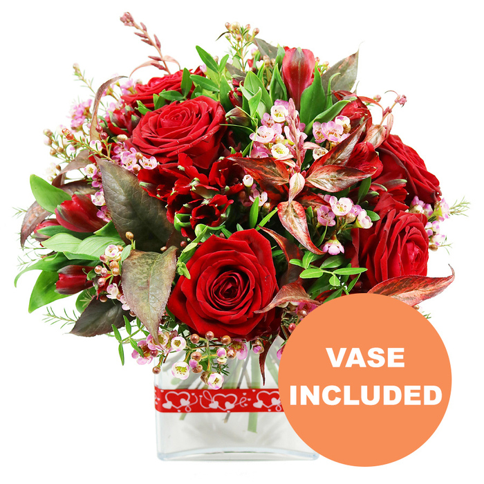 A rich, romantic bouquet in a clear glass vase is photographed against a bright white background, its deep red blooms and fresh greenery arranged with the practiced hand of a local florist familiar with the Brent Cross area. At the heart of the design, large red roses with velvety petals command attention, some tightly closed with a sense of quiet promise, others fully open and luxuriant, displaying their complex swirls and layered forms. Gathered around them are smaller, deep crimson flowers reminiscent of alstroemeria or similar varieties, their multiple, slightly flared petals providing structure, interest, and a lush fullness that fills the dome-like shape of the bouquet. Woven delicately among the larger flowers are fine stems of pale pink and white filler blooms, such as waxflower or limonium, whose tiny clustered blossoms bring a soft, airy quality and gentle contrast against the intensity of the reds. The arrangement is framed by an abundance of green foliage, including broad, solid leaves and more delicate, lighter sprigs, creating a rounded, balanced outline that feels both generous and carefully curated. Standing out among the greens are dramatic leaves in burgundy and reddish-brown tones, some gently veined, adding depth, warmth, and a sophisticated echo of the rose tones. The stems are gathered and displayed in a sleek rectangular glass vase, its minimalist form and transparent sides allowing the clean water and straight stems to be seen, lending a contemporary clarity to the romantic flowers above. Wrapped around the vase is a red ribbon patterned with repeating white heart outlines, an understated nod to love and affection, ideal for anniversaries, proposals, or Valentine's surprises delivered to homes around Brent Cross and Cricklewood. In the lower right corner of the image, a prominent circular orange overlay with bold white capital letters spells out VASE INCLUDED, making it clear that this bouquet arrives ready to be placed straight onto a table, desk, or windowsill.
