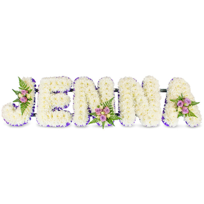 A handcrafted, name-shaped floral tribute reads JENNA in bold, three-dimensional white letters, each letter densely covered in small creamy chrysanthemums that form a plush, cloud-like surface. The flowers' tight petals create a soft, tactile appearance and a delicate fragrance that rises subtly from the composition. A deep purple trim runs along the inner edges of every letter, a crisp line of colour that emphasizes the letterforms and adds a measured grace to the predominantly white design. Three small but intentional floral clusters enhance the tribute: one nests on the upper curve of the J, another settles at the centre of the second N, and a final arrangement crowns the right shoulder of the A. Those clusters combine blush-pink and lavender spray roses, a scattering of pale cream roses, and wisps of dark-green fern to introduce gentle colour variation and leafy texture. Narrow dark structural bars are visible at the base, connecting the letters into a secure composition ready for placement at a funeral service or memorial display. The lighting in the image is soft and even, lending the white chrysanthemums a warm, almost luminous quality while the purple trim provides a quiet focal point. As crafted by Brent Cross Flowers for families who know the local chapels and cemeteries around Brent Cross Shopping Centre, Cricklewood and nearby Golders Green, this arrangement is designed to balance dignity with intimacy-an elegant, personal way to honour a loved one's memory with floral craftsmanship and heartfelt attention.