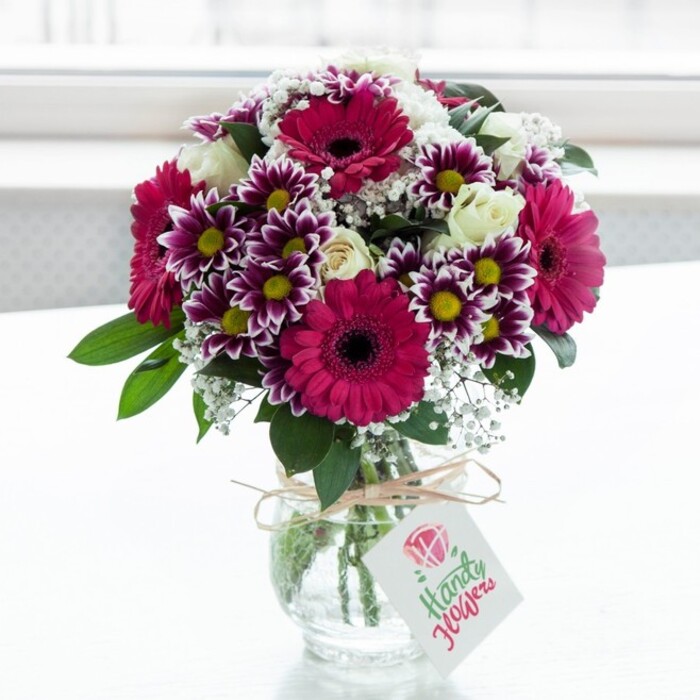 A lush, joyful floral arrangement is captured in a bright, minimalist scene, as though just placed on a windowsill in a Brent Cross flat after a same-day delivery. The bouquet spills generously from a clear, textured glass vase with a wide mouth, the stems neatly gathered and visible through the water. At the heart of the design are bold gerbera daisies in vivid shades of fuchsia and magenta, their broad, layered petals radiating out from deep, almost chocolate centres that lend a sophisticated contrast. Surrounding these focal blooms are distinctive bicolour chrysanthemums: each daisy-like flower has petals tipped in rich purple that gently fade to white toward the centre, encircling bright yellow eyes that glow warmly in the soft light. Between these statement flowers, clusters of creamy white spray roses are carefully placed, some just beginning to open, their tighter buds adding a sense of anticipation and longevity to the display. Fine, cloud-like sprays of white baby's breath weave through the arrangement, their tiny blossoms creating a delicate mist that softens the strong colours and adds visual lightness. Glossy, dark green foliage emerges from the base and peeks between blooms, providing structure and a fresh, garden-picked feeling that a local florist might gather from markets near Golders Green. The vase is finished with a simple, natural twine bow tied around its neck, from which a small, rectangular white gift tag dangles, decorated with a stylised pink rose emblem and elegantly flowing script, hinting at a personal note tucked into a flower delivery. The softly diffused white background ensures there are no distractions, allowing the intricate textures, vibrant hues, and careful craftsmanship of the bouquet to shine, making it ideal for marking birthdays, anniversaries, or heartfelt thank-yous across Brent Cross and the surrounding North London neighbourhoods.