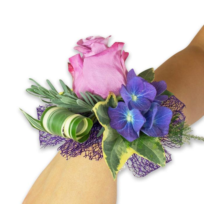 Displayed on a pale forearm against a clean white backdrop, this wrist corsage reads as a sophisticated little garden. The central bloom is a generous pink rose, partially open so its petals reveal a soft gradient from a deeper fuchsia at the ruffled edges into a lighter blush toward the heart; the petal surfaces have a velvety finish that hints at a delicate fragrance. Nestled around this focal rose are three dense clusters of deep indigo-purple hydrangea, each tiny flowerlet bearing a luminous blue-white center that lends complex texture and depth. Interwoven foliage includes long, narrow silvery-green sprigs that resemble eucalyptus, giving a gentle, cooling contrast, while glossy variegated ivy leaves with dark green centers edged in pale cream bring a touch of traditional elegance. A boldly striped broad leaf, white and green, is skillfully curled into a tight spiral to provide modern sculptural interest, and beneath the florals a sheet of open-weave dark purple mesh peeks out, tying the composition together and amplifying the hydrangea tones. The result is a balanced mix of classic romance and contemporary design that feels both tactile and refined-the silky rose, crisp ivy, and feathery greens working together as if arranged on a bench in Brent Cross Gardens on a quiet spring morning. Prepared by Brent Cross Flowers with an eye for formal occasions, this corsage is ideal for proms, weddings, or graduations and is crafted to be easily worn and photographed among local venues and streets near the shopping centre.