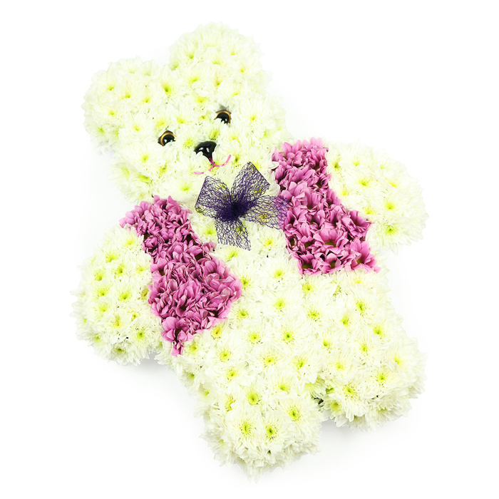 In this image, a teddy bear created completely from flowers appears almost life-like in its tenderness, yet remains clearly a work of careful floristry. The bear sits upright against a spotless white background, its form sculpted from a carpet of crisp white chrysanthemums that cover every visible surface. Each bloom is snugly set next to its neighbour, following the gentle curves of the bear's head, rounded snout, ears, and compact limbs, which are tucked in slightly as though the bear is resting patiently. The chrysanthemums' fine petals and soft texture give the impression of a fluffy, comforting coat. The facial features are modest but full of personality: two dark, bead-like eyes look outward with a calm, reassuring gaze, while a small triangular black nose anchors the centre of the face. Beneath it, a faint curve of soft pink detail suggests a delicate, almost whispered smile, lending a sense of warmth and affection. Across the chest, a waistcoat of rich pink and magenta chrysanthemums provides a beautiful contrast, the flowers arranged in two defined sections that line up like neatly pressed lapels. Just where the vest meets the neck, a bow made of light, gauzy purple netting rests gently on the petals, its airy loops introducing a sense of lightness and grace. The overall effect, enhanced by the clean lighting and absence of distractions, is one of quiet comfort and innocence. Families in Brent Cross often choose this style of floral teddy bear for funerals or memorials near the North Circular or local synagogues and churches, especially when remembering a child or someone whose memory is held with particular tenderness.