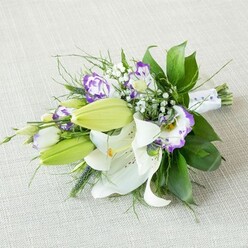 A long, elegant bouquet of lilies and lisianthus rests at a gentle angle on a pale beige, textured fabric, like a bridal arrangement being admired before the ceremony in a quiet Brent Cross sitting room. The composition feels both classic and contemporary, built around large white lilies whose smooth petals arch open to reveal golden-yellow stamens and fine reddish-brown flecks dusted near the throat of each bloom. Their fragrance is almost imaginable, that familiar, soothing lily scent that often fills homes off the North Circular and nearby streets. Several bright green lily buds are tucked along the length of the bouquet, their tightly closed tips adding height, freshness, and the suggestion of tomorrow's blossoms. Between the lilies weave clusters of lisianthus, some in soft white kissed with delicate lavender, others edged in a deeper royal purple, their ruffled petals giving the overall design a romantic, slightly whimsical feel. Sprays of tiny white baby's breath dot the arrangement, creating a misty veil that bridges the transition between flowers and foliage. The greenery forms a rich frame, combining broad, shiny leaves with finer, spikier stems that flick outward and add a sense of natural movement, like a breeze drifting through a garden near Clitterhouse Playing Fields. At the base, the stems are bound tightly with a crisp white satin ribbon, subtly decorated with small pearl-like beads that catch the light along the narrow end of the bouquet, implying it is meant to be held or presented with ceremony. The neutral fabric below and the soft, even light above keep the focus entirely on the interplay of white, green, and purple, creating an impression of quiet joy and refined celebration, perfect for a wedding, milestone birthday, or thoughtful same-day delivery around Brent Cross.